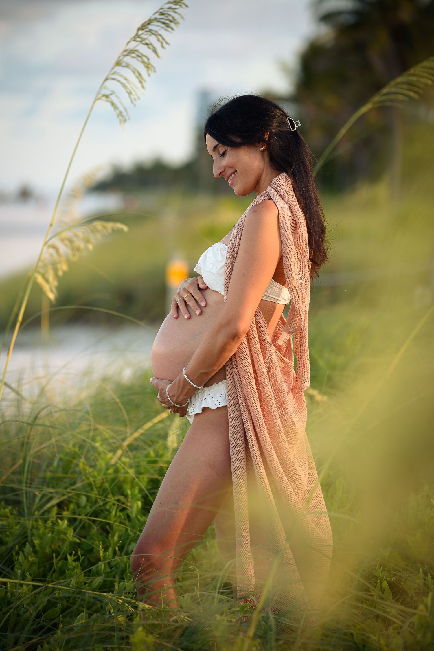 Smiling pregnant woman in pink wrap and white lace outfit cradling her baby bump outdoors in lush green coastal grass.