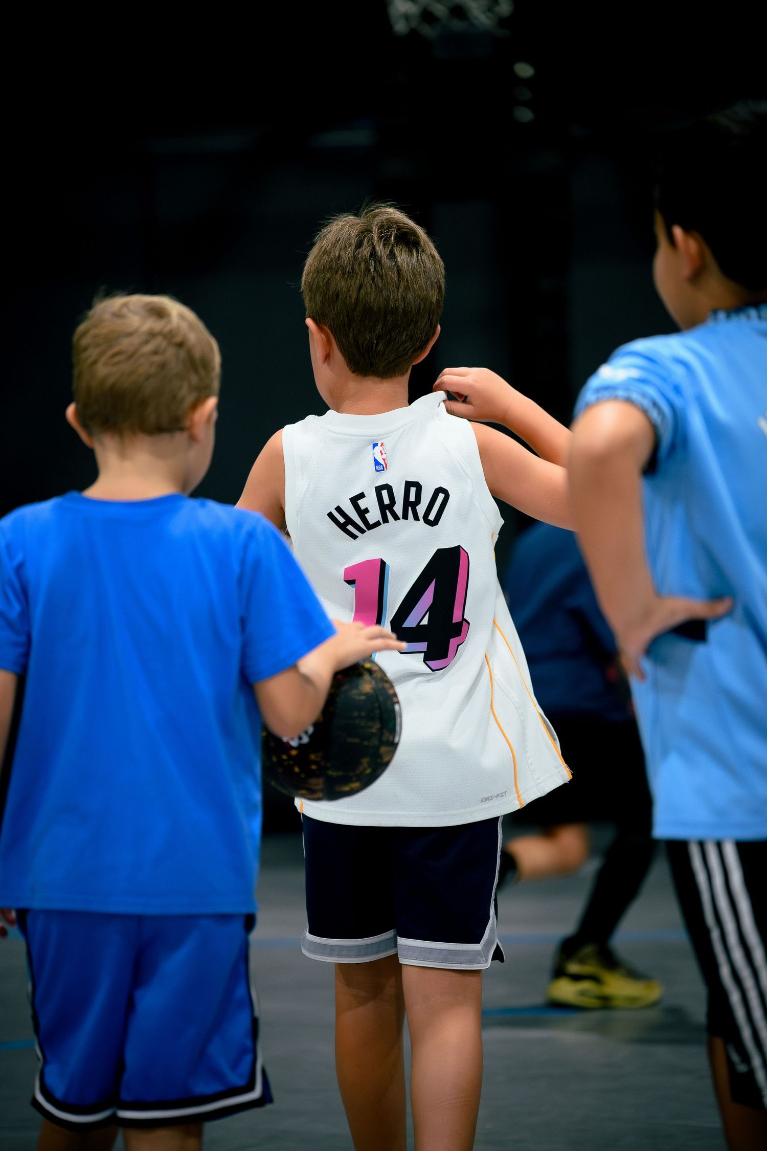 Young boy wearing Miami Heat Tyler Herro number 14 jersey playing basketball with friends indoors.