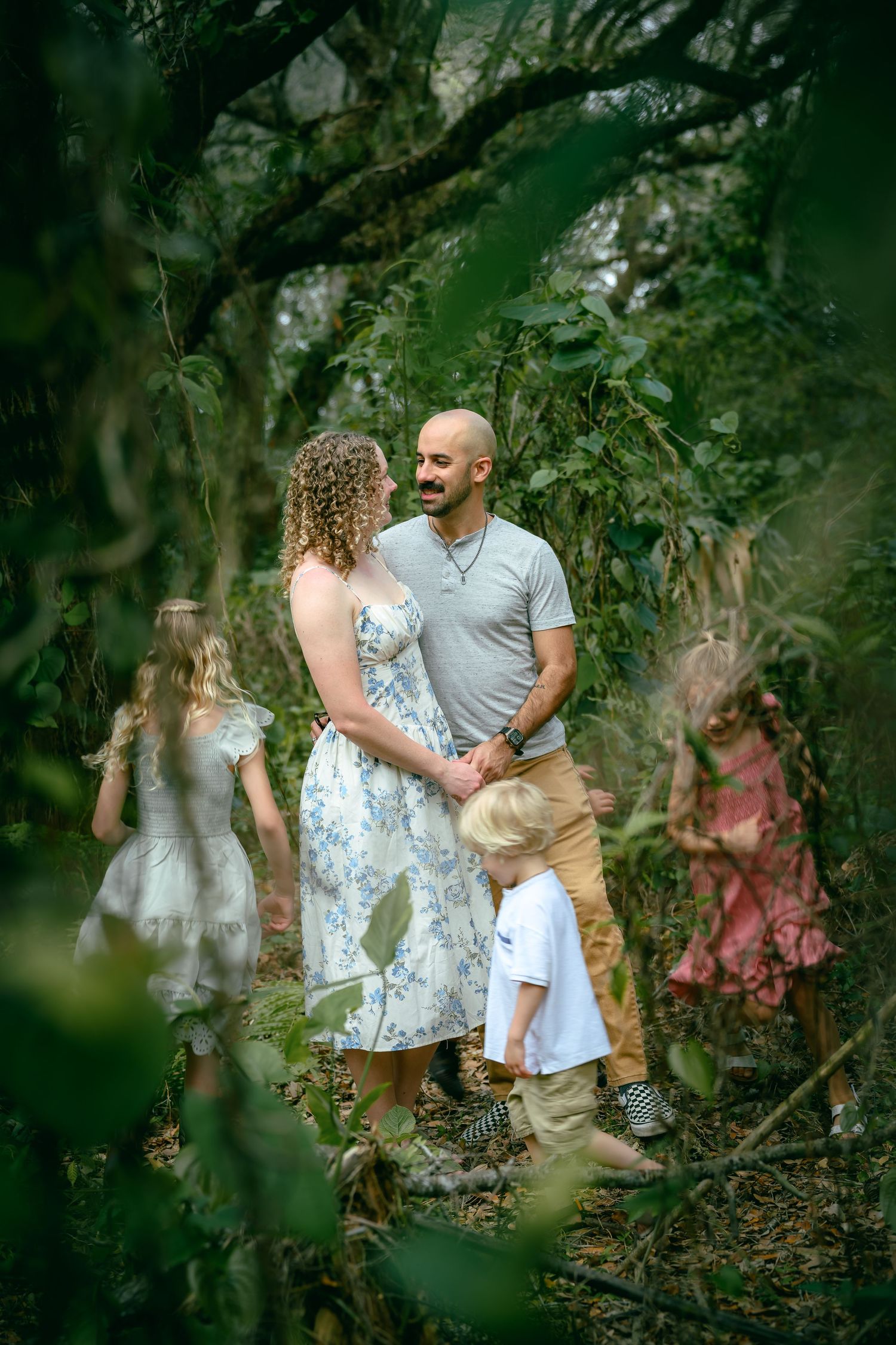 A family of five stands together in a lush green forest, parents gazing lovingly at each other while children play around them.