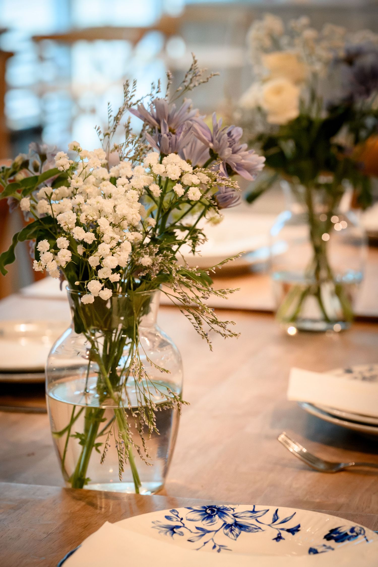 White floral centerpieces in glass vases adorn a wooden table with blue accents at an elegant reception setting.