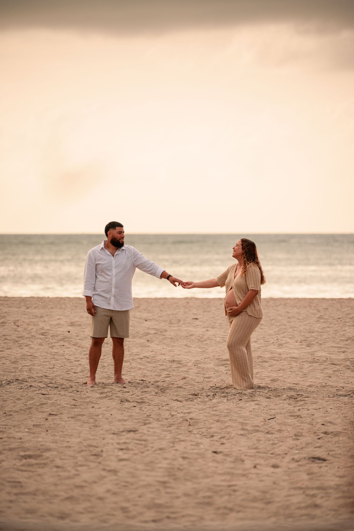 Couple holding hands on a sandy beach at sunset, woman appears pregnant, ocean waves in background.