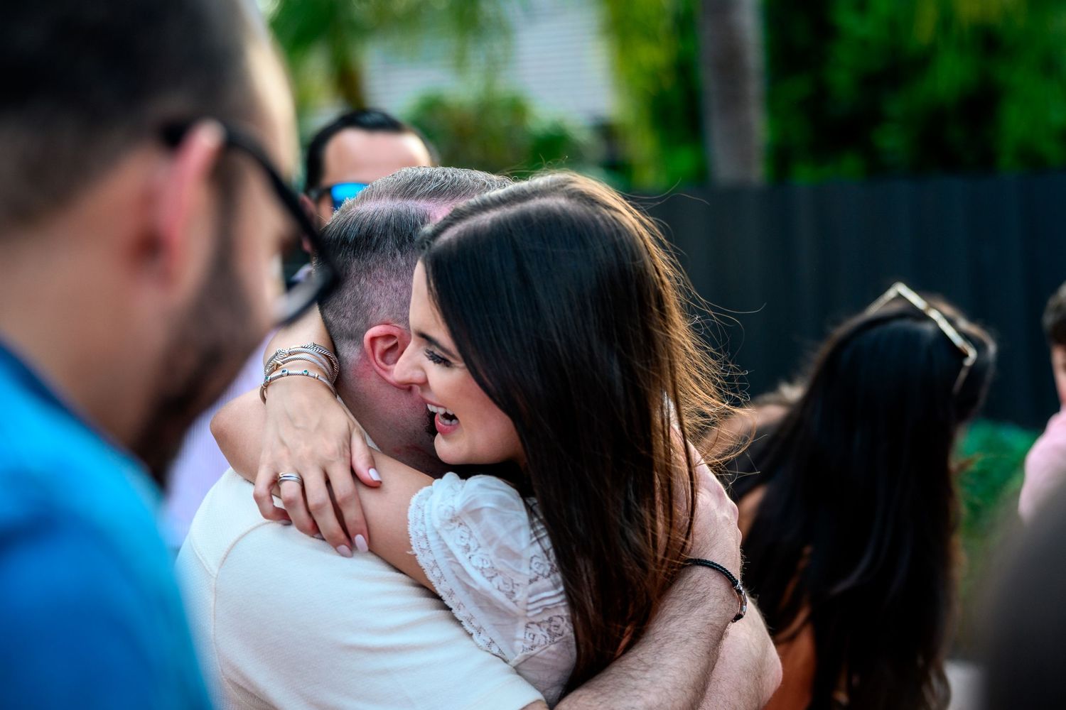 Two people share a heartfelt embrace at an outdoor social gathering surrounded by guests.