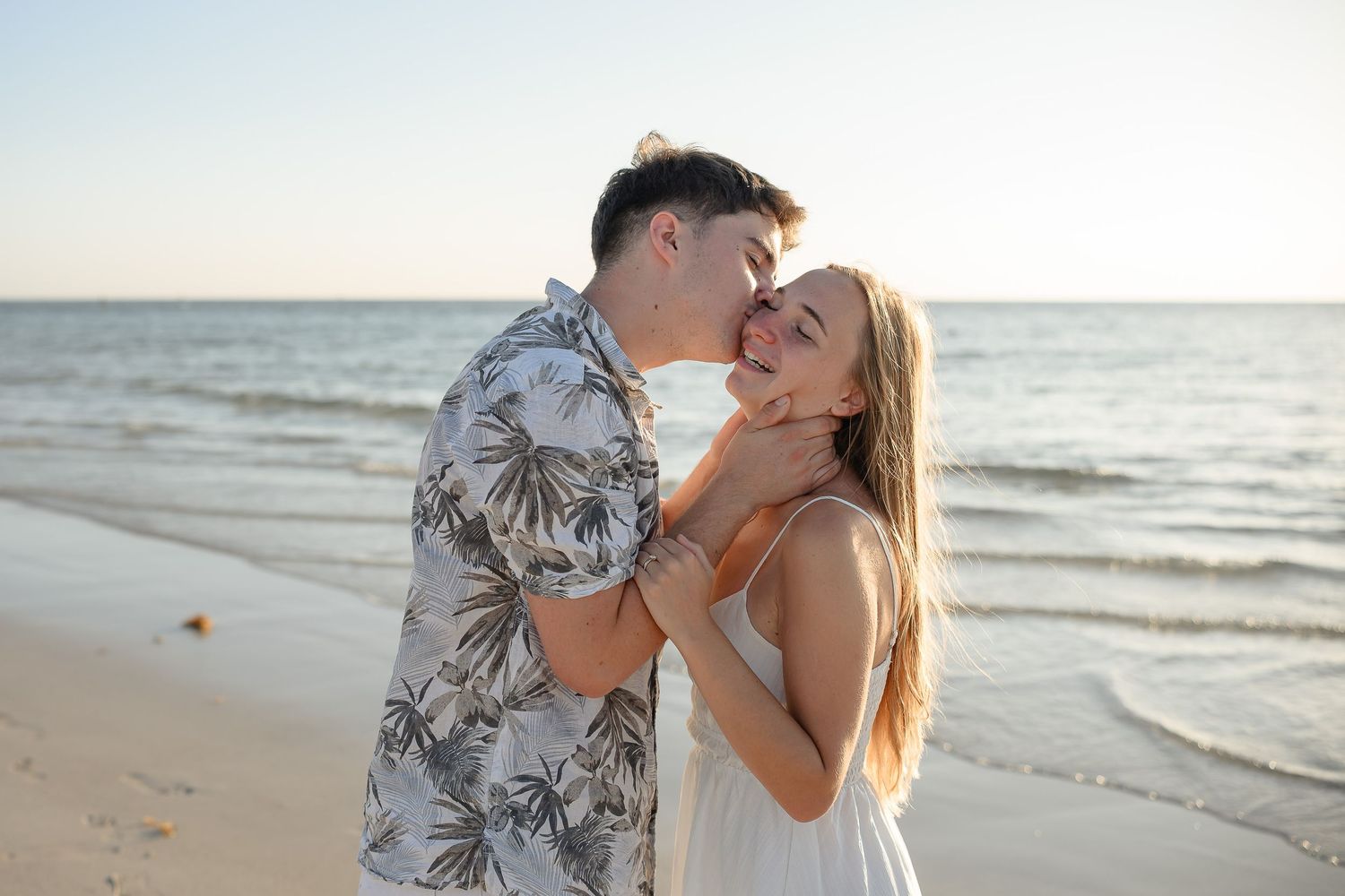 Couple sharing a romantic moment on a sandy beach at sunset, man kissing woman's cheek as she smiles.
