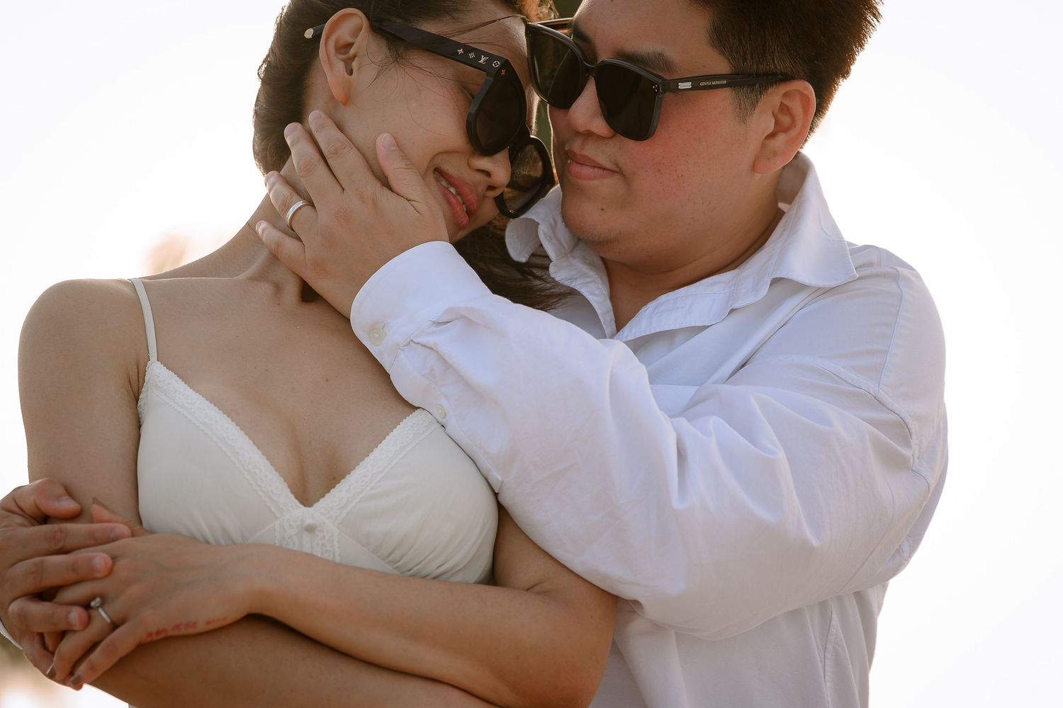 A romantic couple embracing outdoors, both wearing sunglasses, woman in white dress, man in white shirt, in warm sunlight.