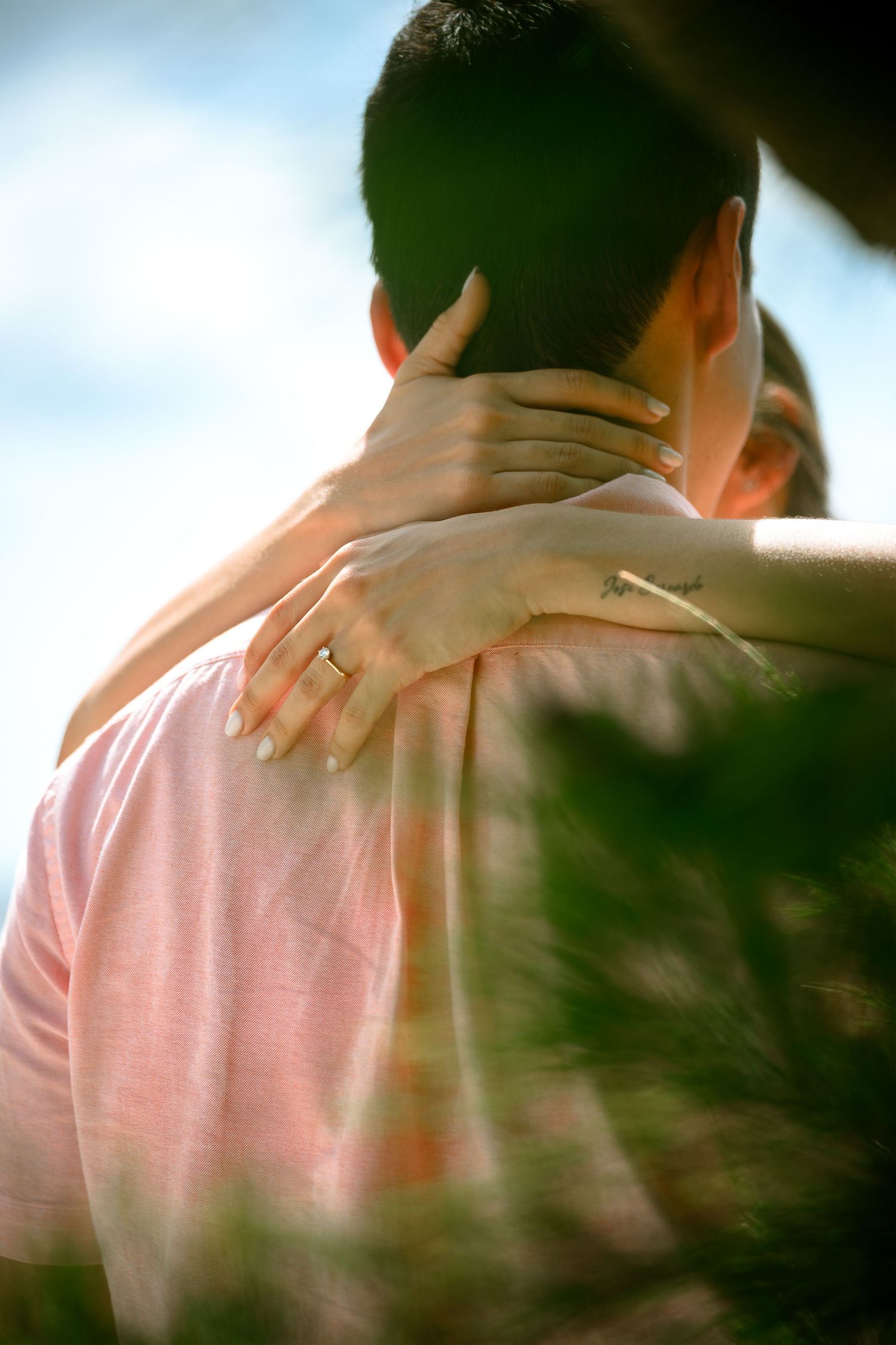 A couple embraces outdoors, woman's hand with engagement ring resting on man's shoulder in soft natural light.