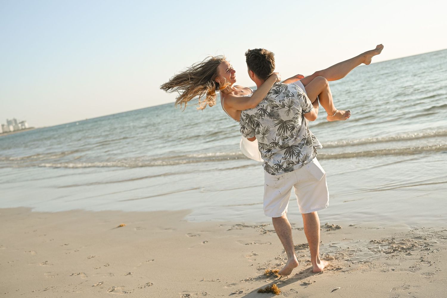 A happy couple playfully spinning on a sunny beach with ocean waves in the background.