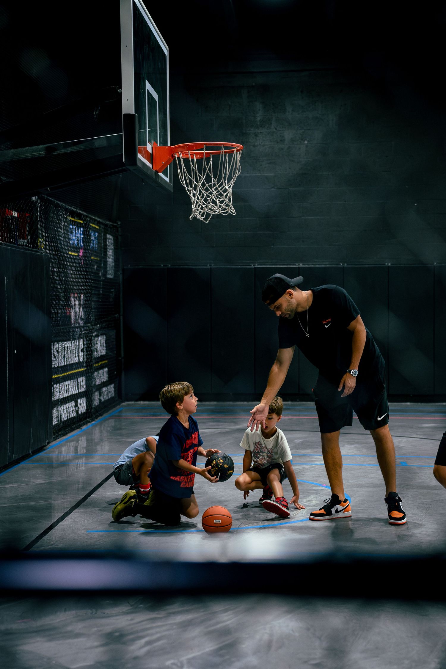 A basketball coach instructs two young kids on an indoor court beneath a hoop during a training session.