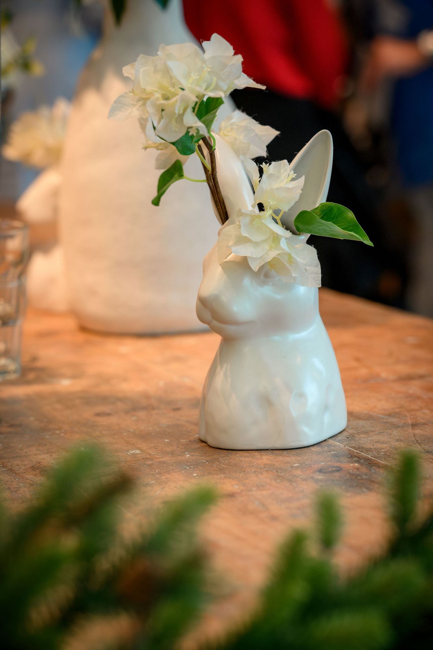 White ceramic vase with delicate white flowers arranged on a textured surface with greenery in the foreground.