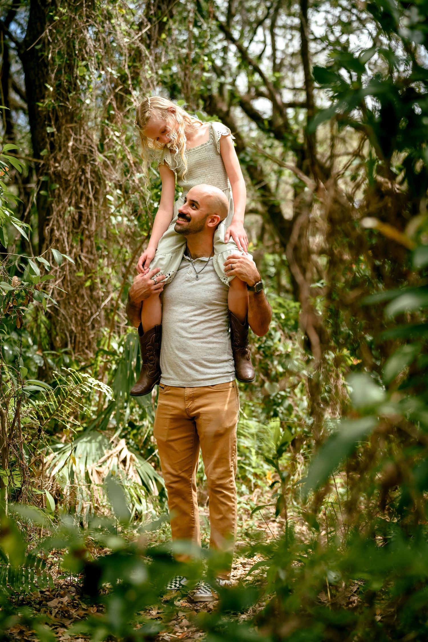 A smiling bald man carries a young blonde girl on his shoulders while exploring a lush green forest trail.