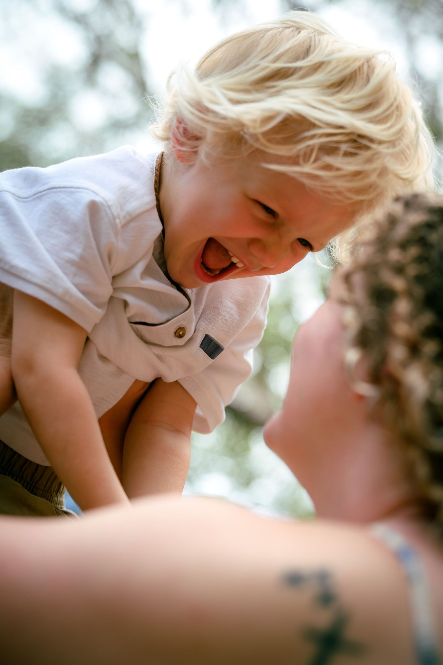 A laughing blonde toddler is lifted up by a curly-haired woman in a joyful outdoor moment.