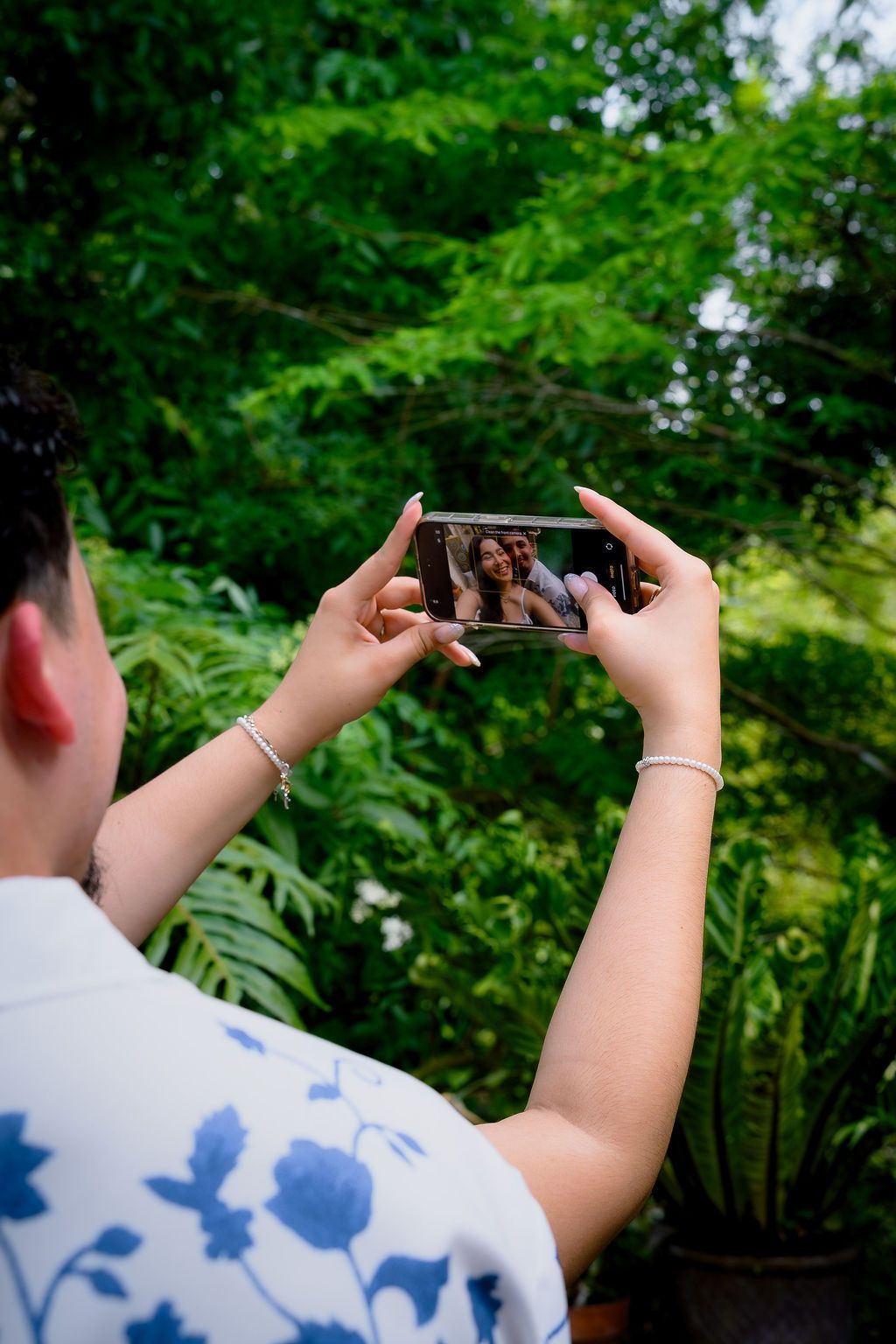 Woman holding smartphone taking a selfie with a friend in a lush green garden setting.