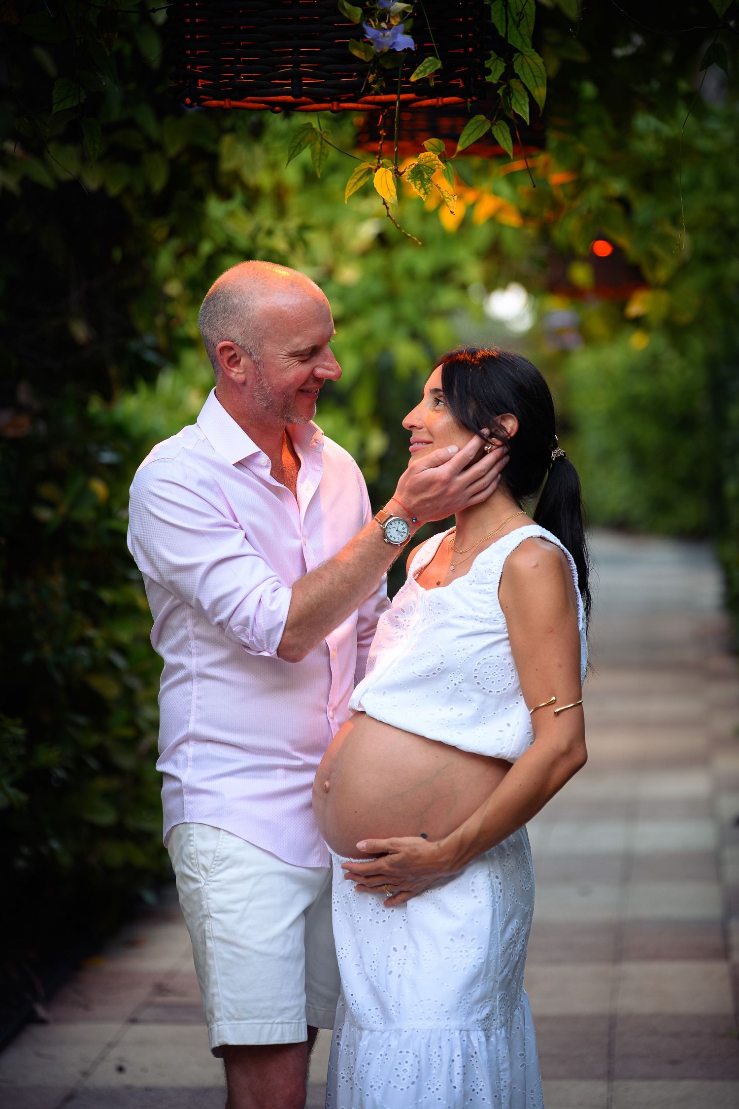A bald man tenderly cups a pregnant woman's face outdoors, both smiling warmly amid lush green foliage.
