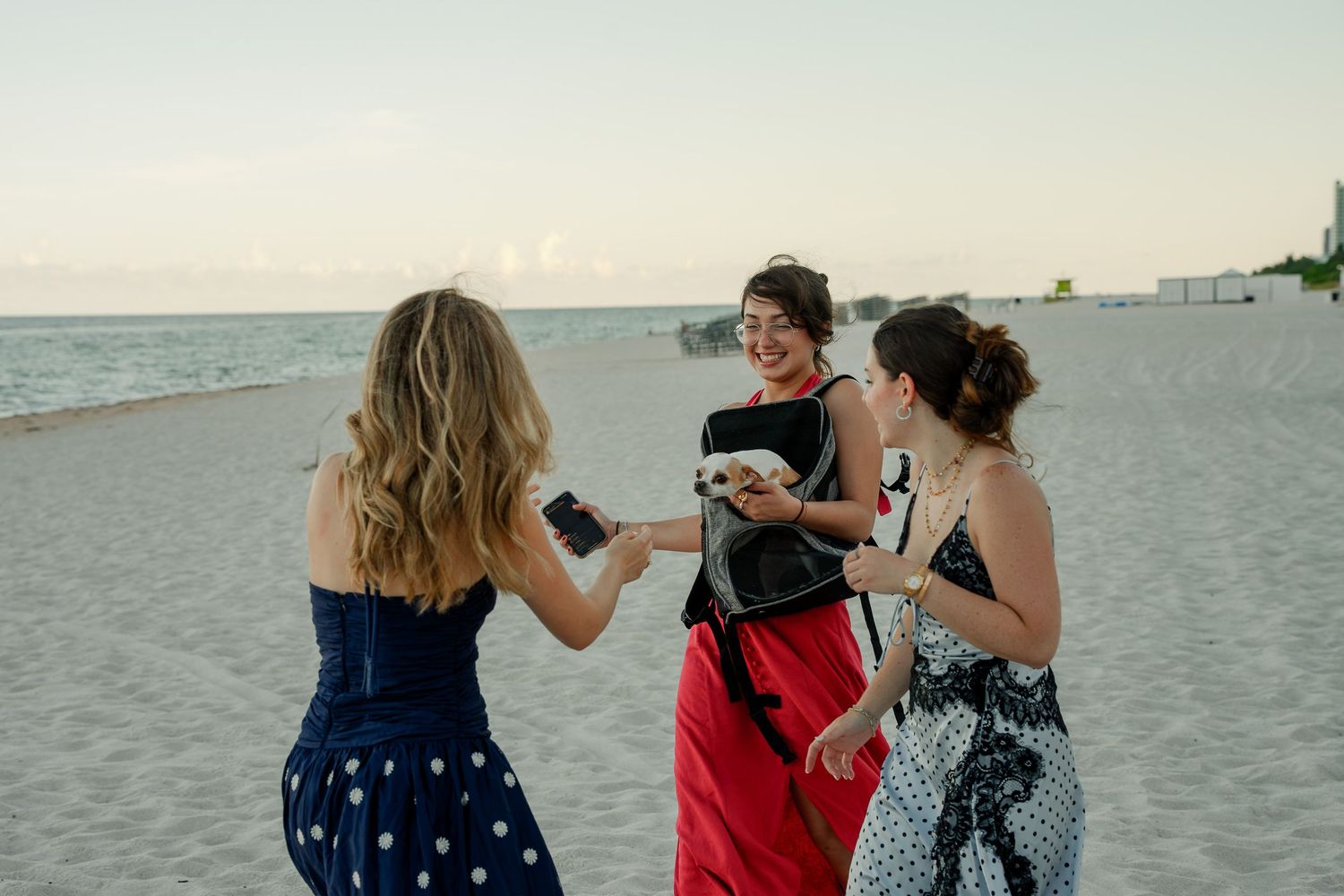 Three women in dresses laughing and sharing food on a sandy beach at sunset.