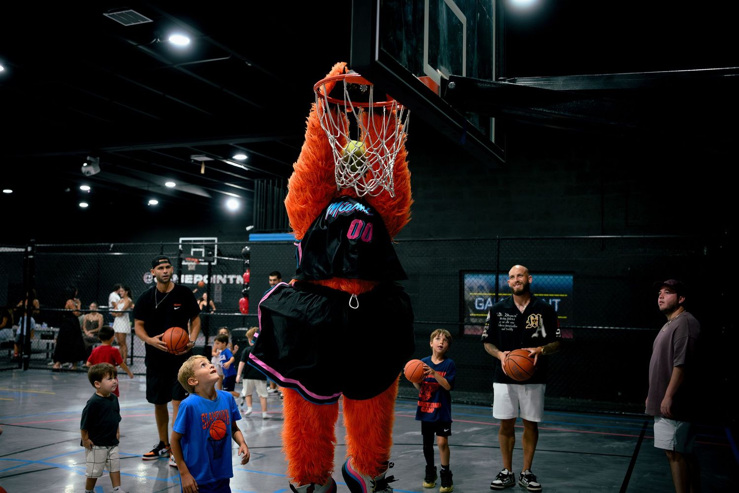 Orange furry mascot dunking basketball at indoor court while children watch during a fun sports event.