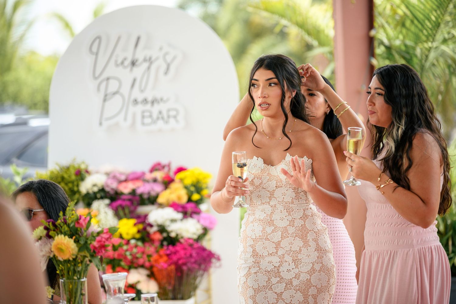 Bridesmaids in pink dresses gather around a bride in white during an outdoor wedding ceremony with tropical flowers.