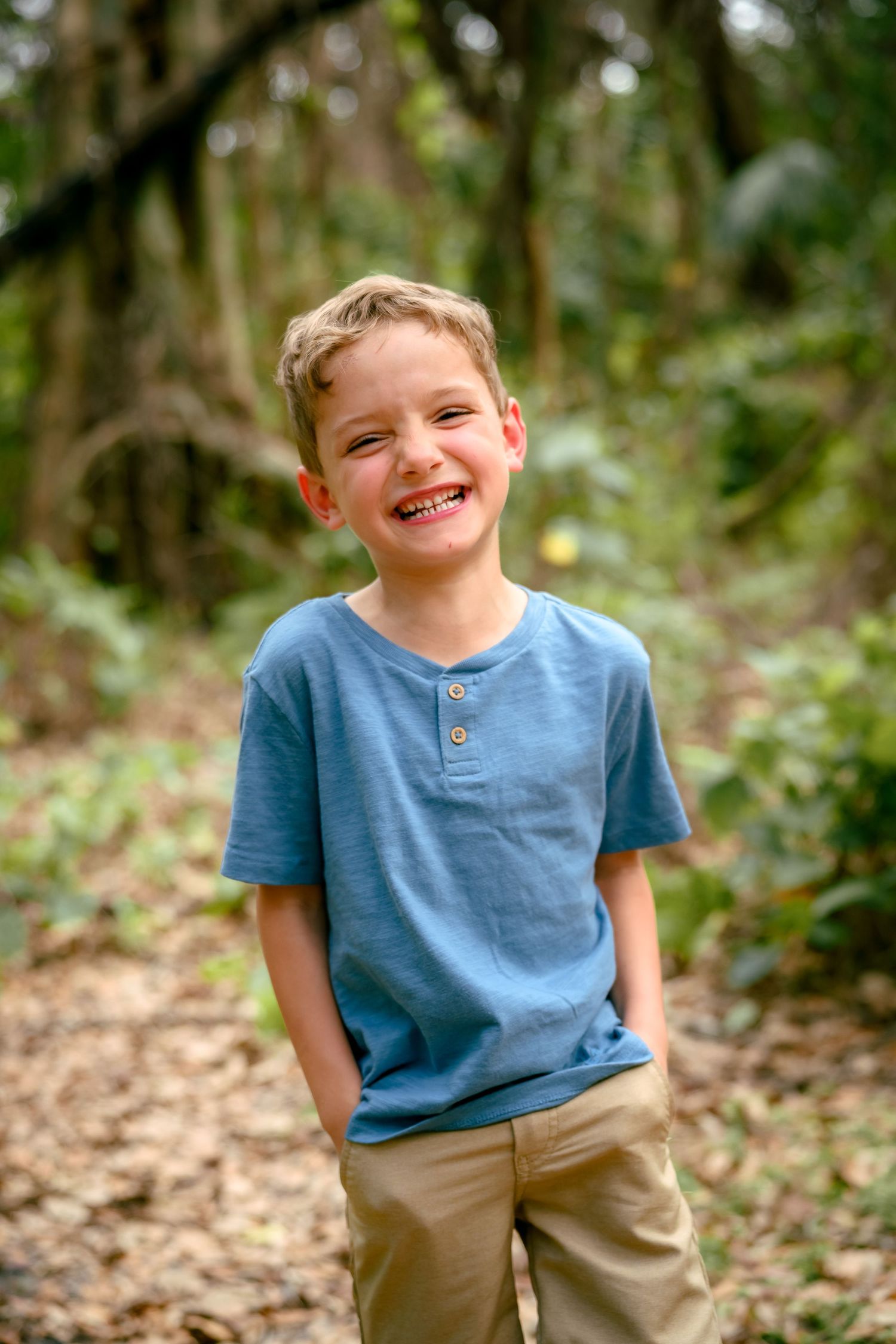 A smiling young boy in a blue henley shirt and khaki shorts stands on a forest trail surrounded by lush green trees.