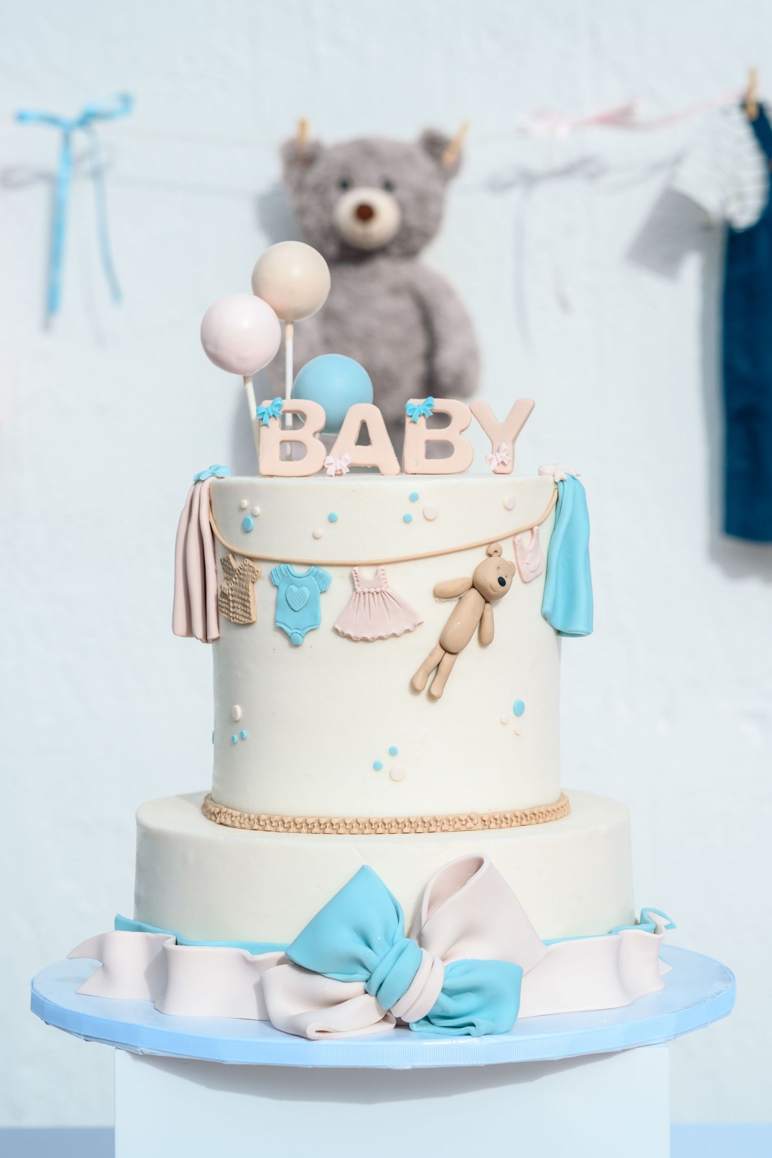 Two-tier baby shower cake with blue bow, teddy bear decorations, and BABY topper on pastel blue background.