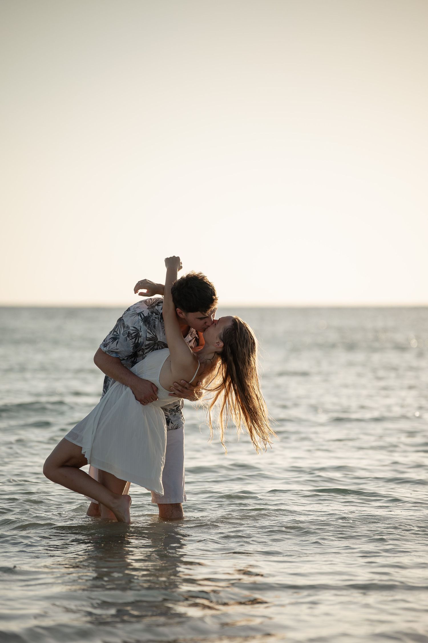 Romantic couple kissing and dancing in shallow ocean waves at sunset on a beautiful beach.