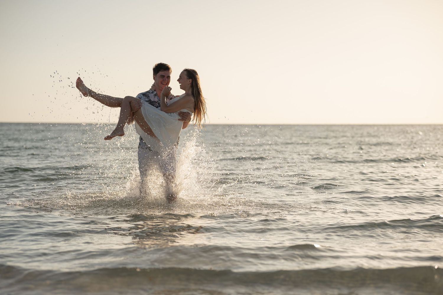 A romantic couple plays in ocean waves at sunset, man carrying woman in his arms on a beautiful beach.