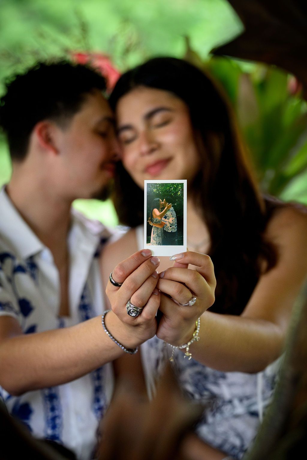Couple sharing a romantic moment while holding a Polaroid photo of themselves in a lush tropical garden setting.