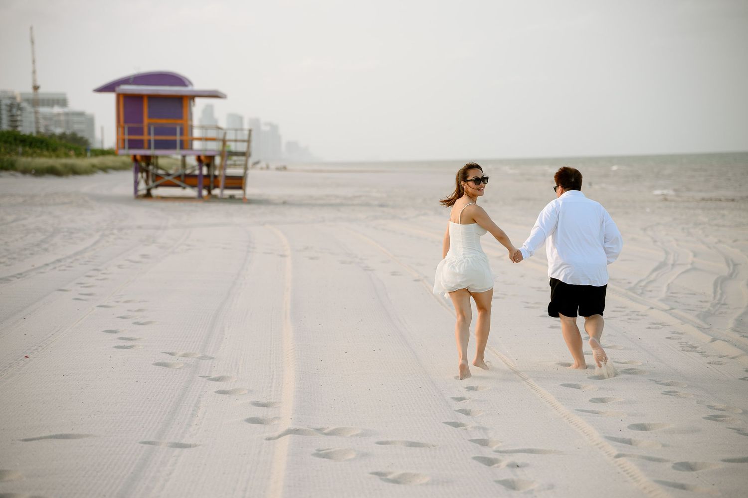 Couple holding hands walking barefoot on a sandy Miami Beach with a colorful lifeguard stand in the background.