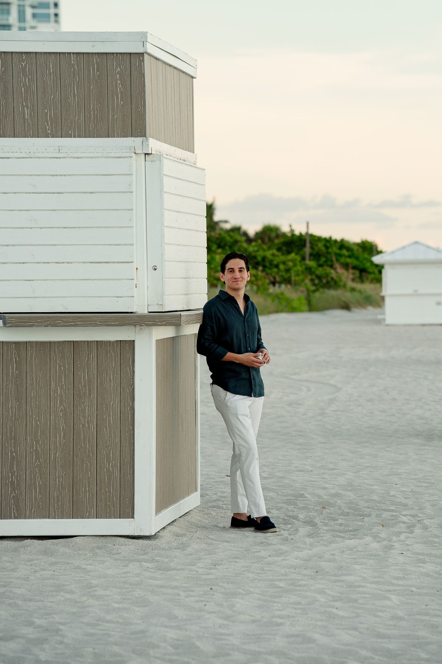 A man in a dark shirt and white pants stands by a beach hut on sandy shore at dusk.