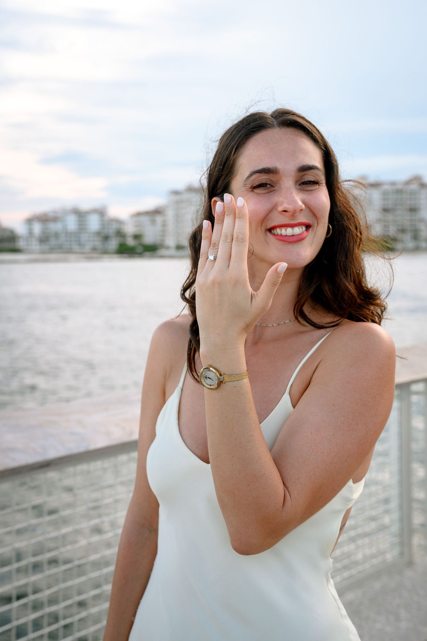 Happy woman in white dress showing engagement ring by waterfront, wearing gold watch, smiling outdoors.