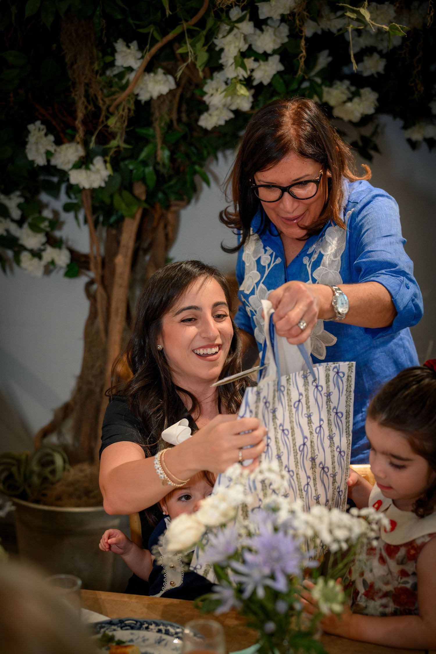 People gather around a table working with flowers and floral arrangements under a blooming tree with white blossoms.