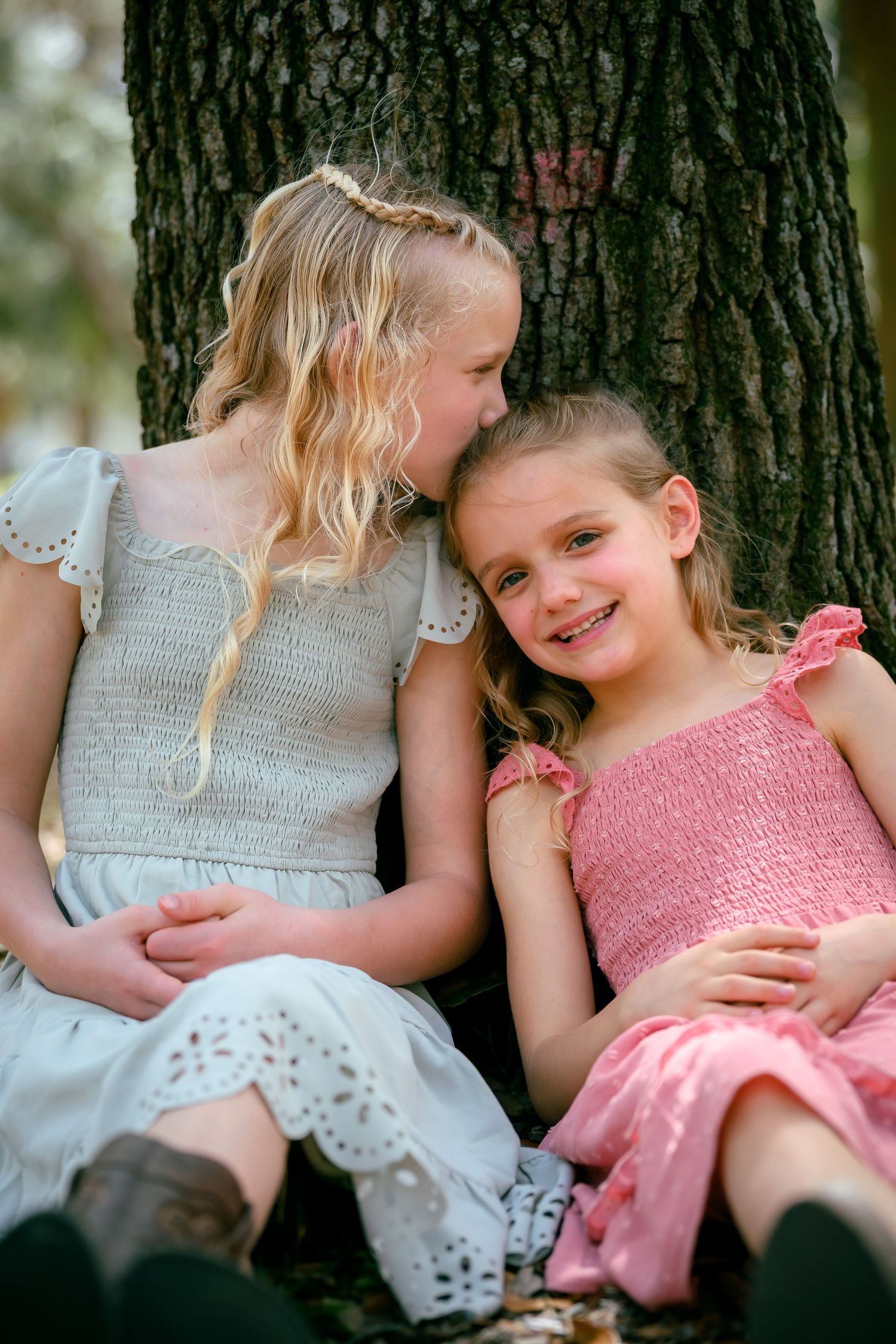 Two young girls sit together by a tree, one whispering to the other who smiles, wearing white and pink dresses.