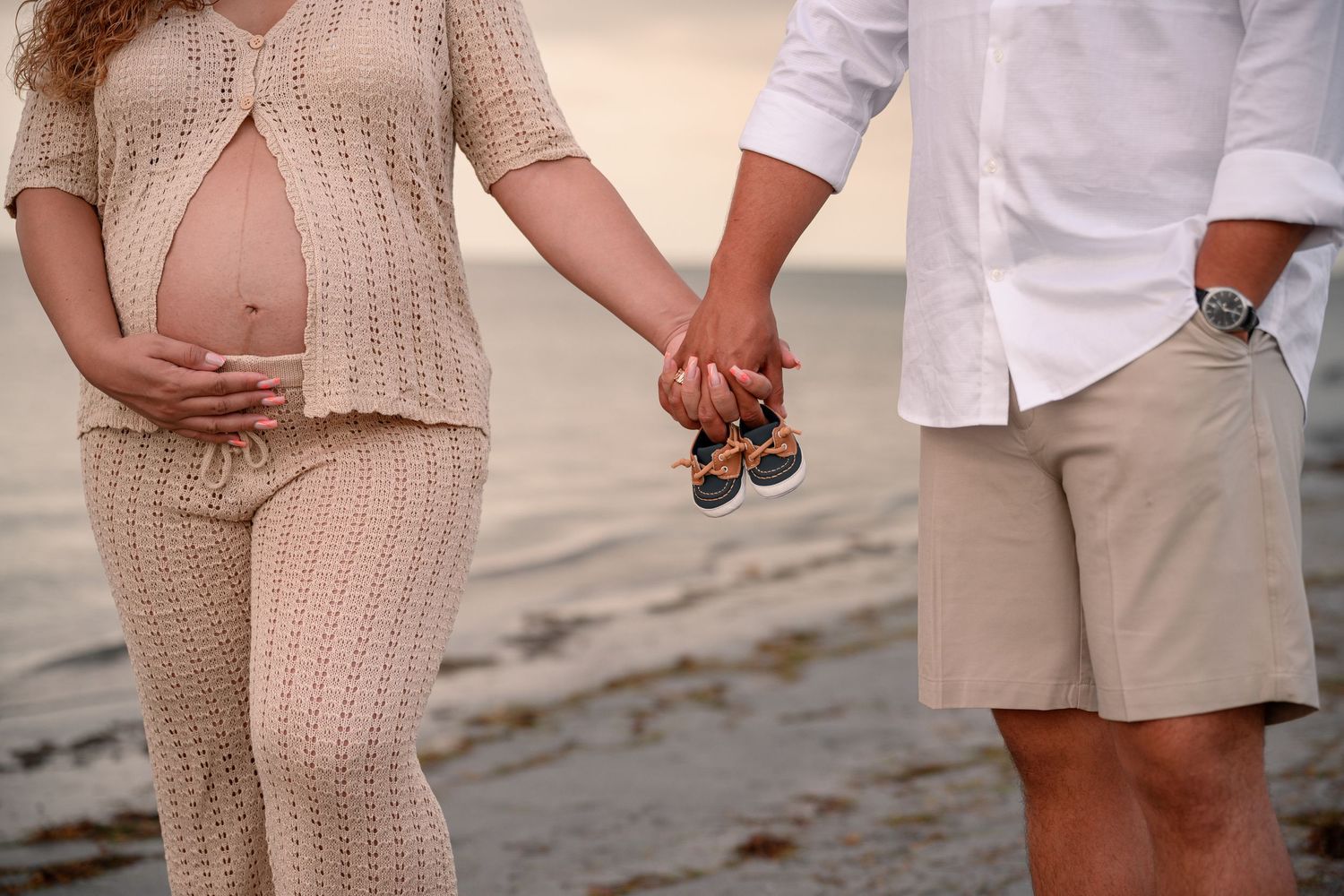 Pregnant couple holding baby shoes on beach, woman in knit outfit cradling bump, romantic maternity photo.