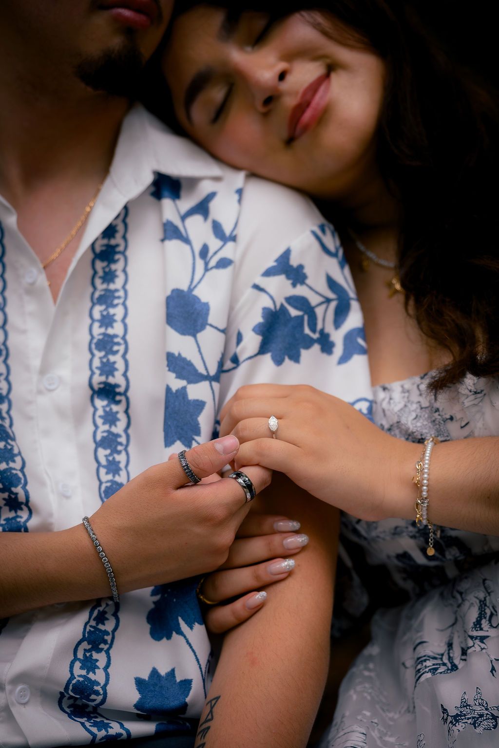 Couple sharing a tender moment, woman resting head on mans chest showing engagement ring, wearing blue floral shirt.