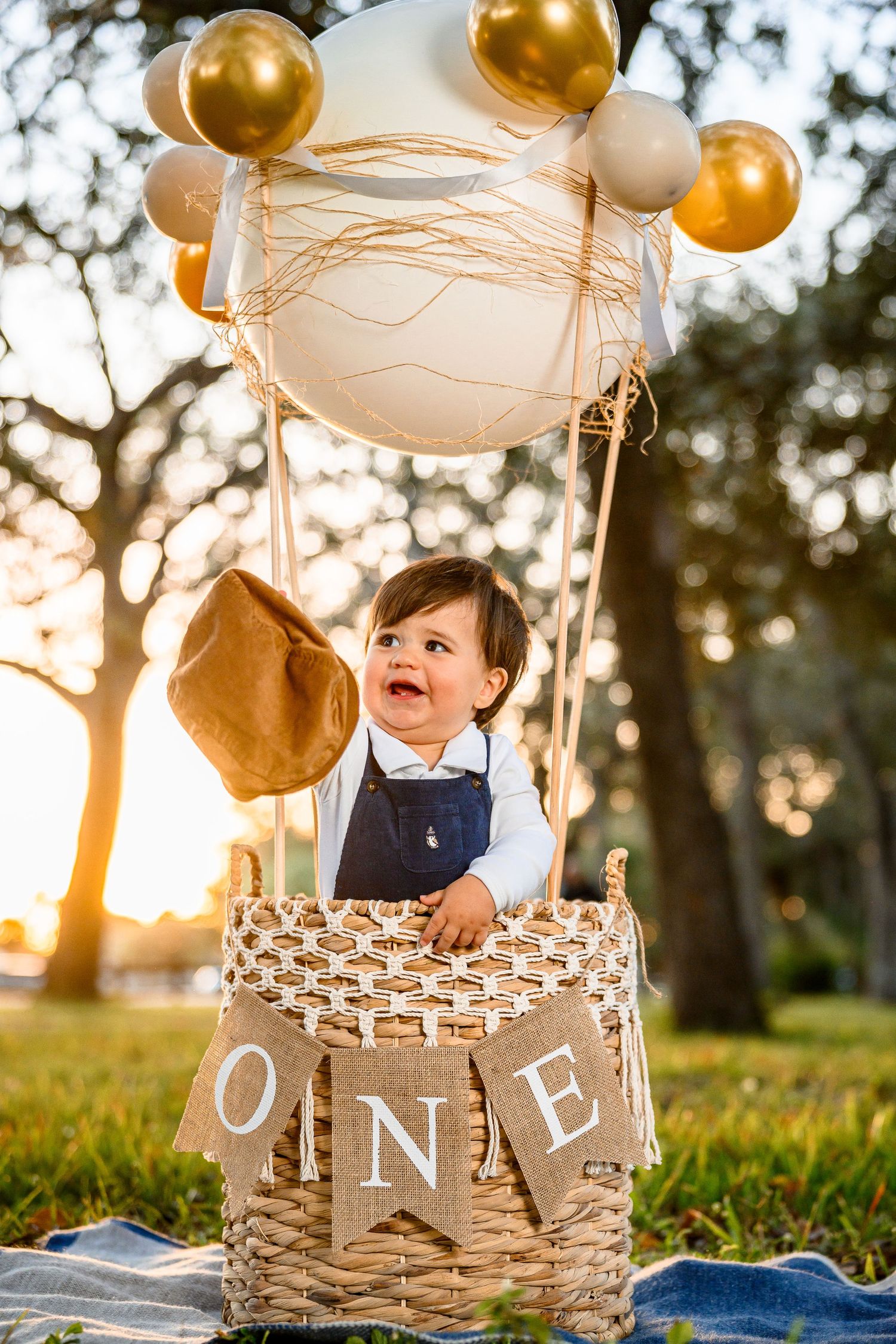 Baby's first birthday photo in a decorative hot air balloon basket with gold and white balloons outdoors.