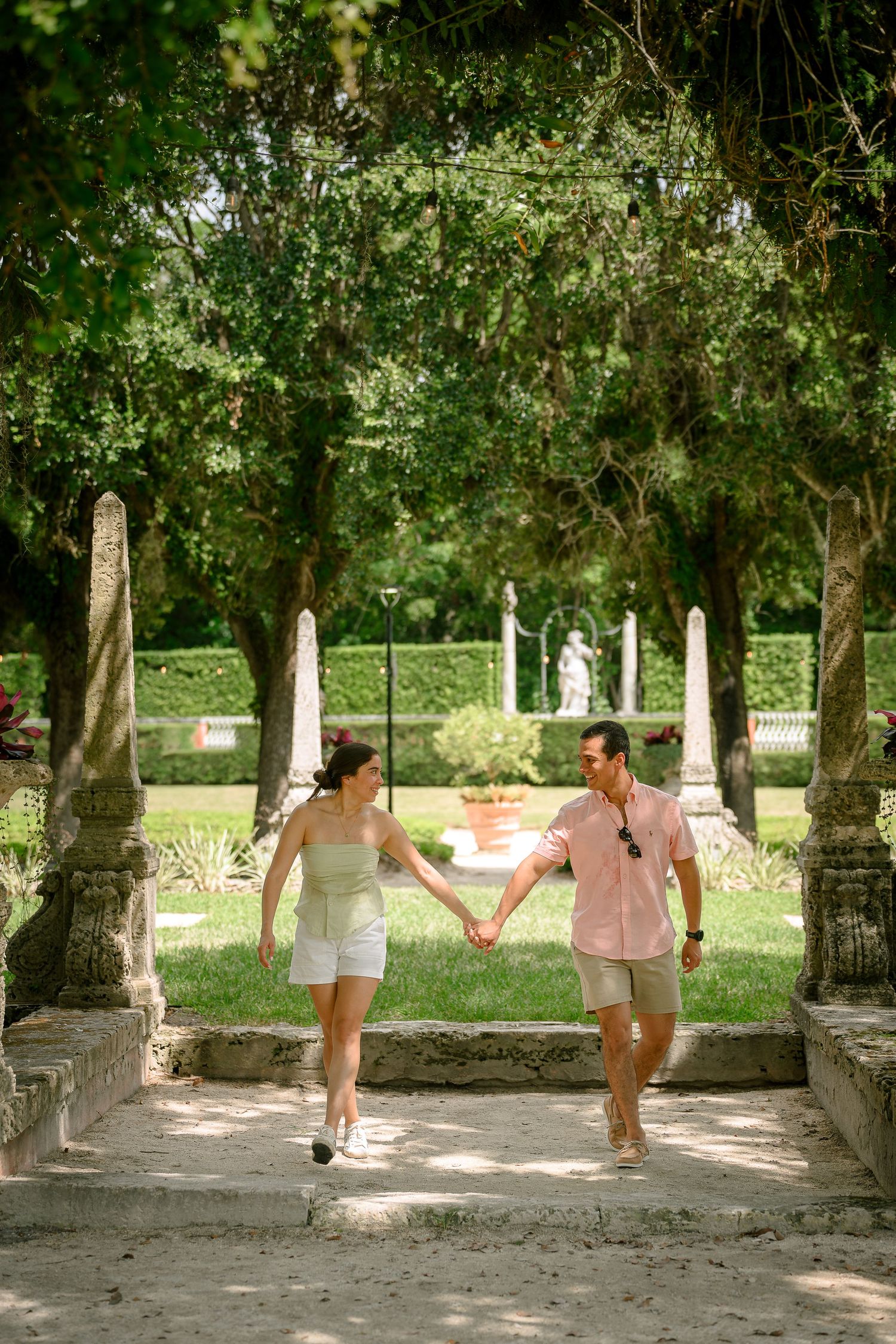 Couple holding hands walking through lush garden archway with stone columns and green trees at Vizcaya Museum.