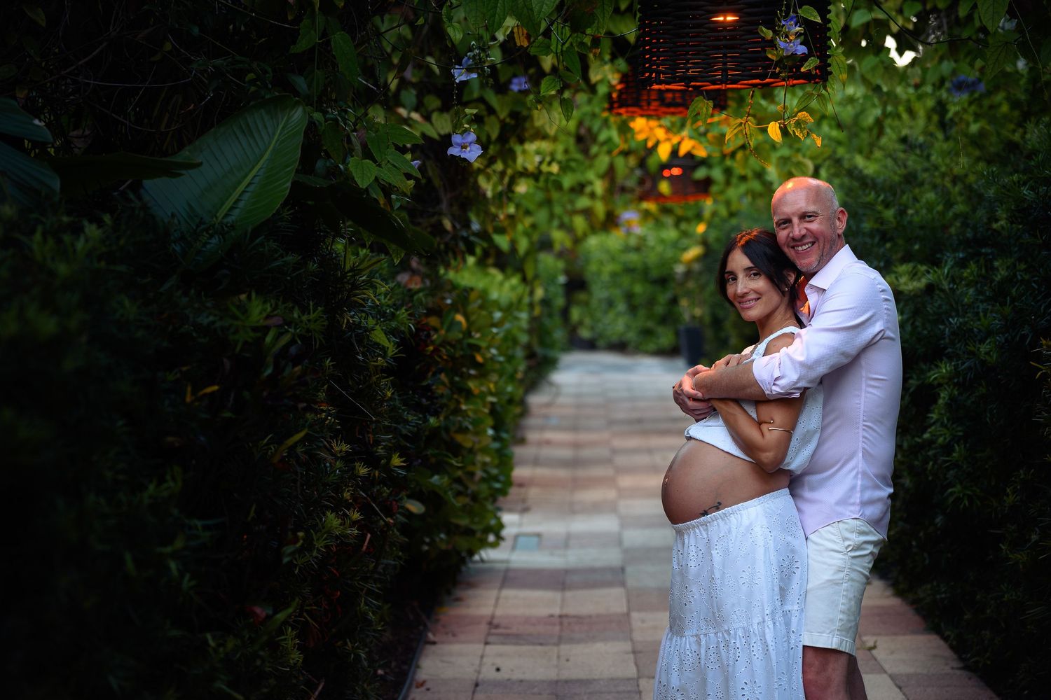 Couple embracing on a lush garden pathway, pregnant woman in white dress with partner in white shirt.