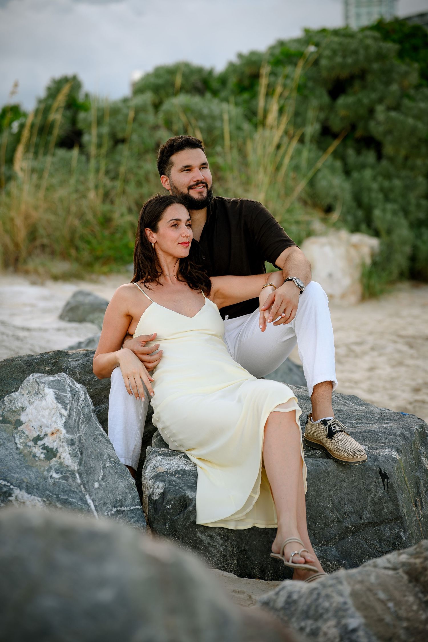 A couple sits on rocks at a beach, woman in yellow dress leaning on man in black shirt and white pants.