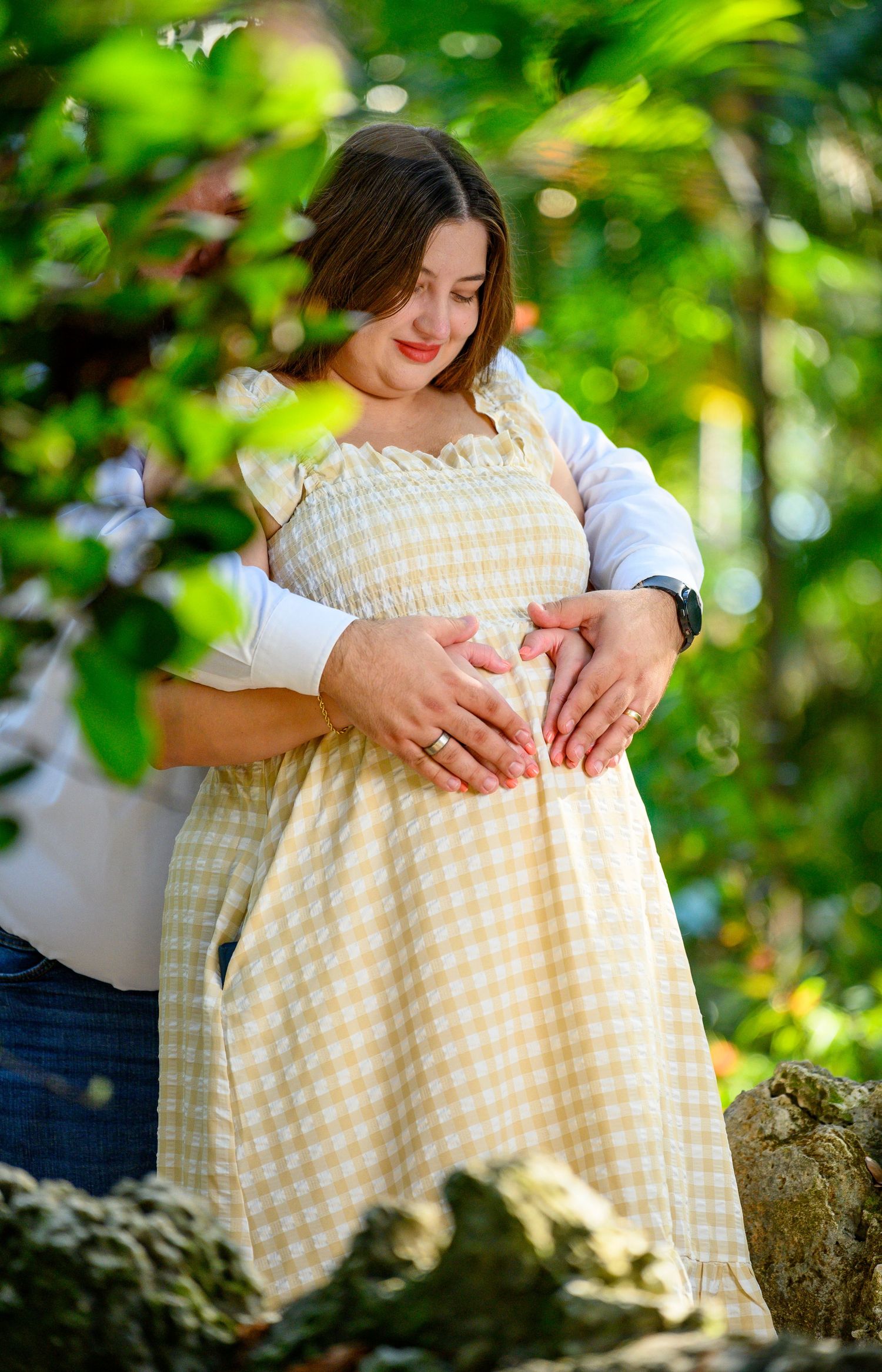 Pregnant woman in yellow gingham dress with partner forming heart shape on baby bump outdoors.