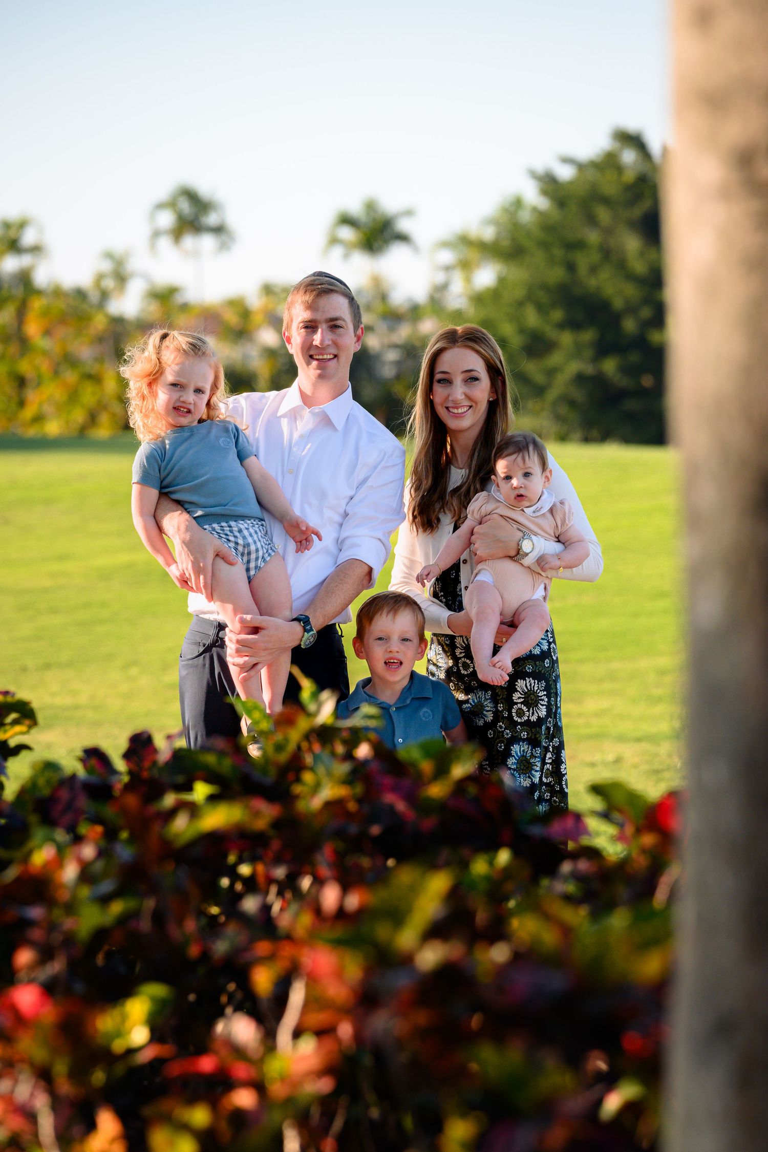Happy family of five posing outdoors on green lawn with tropical palms, parents holding three young children.