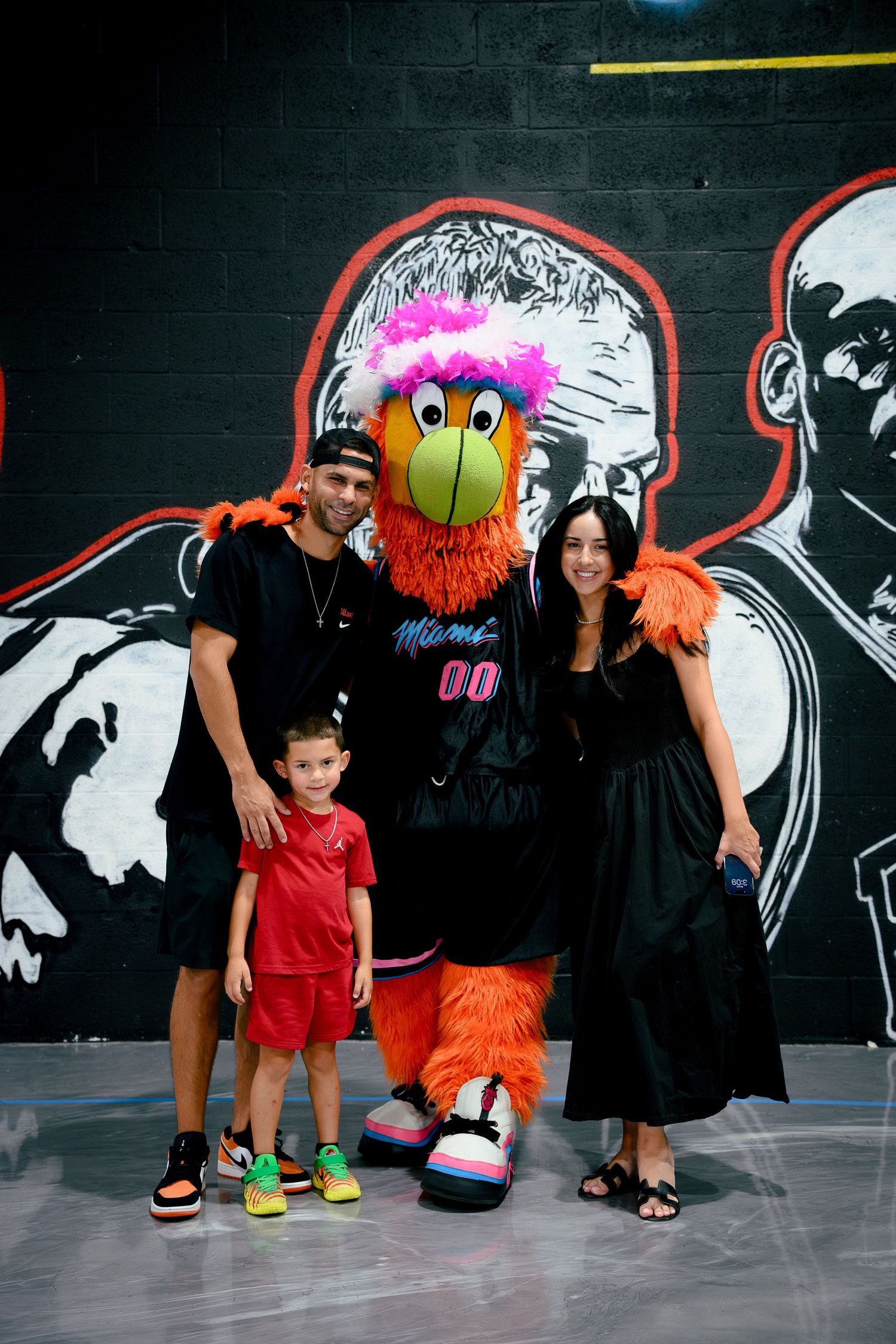 Family posing with Miami Heat mascot Burnie at an NBA arena with player murals in the background.
