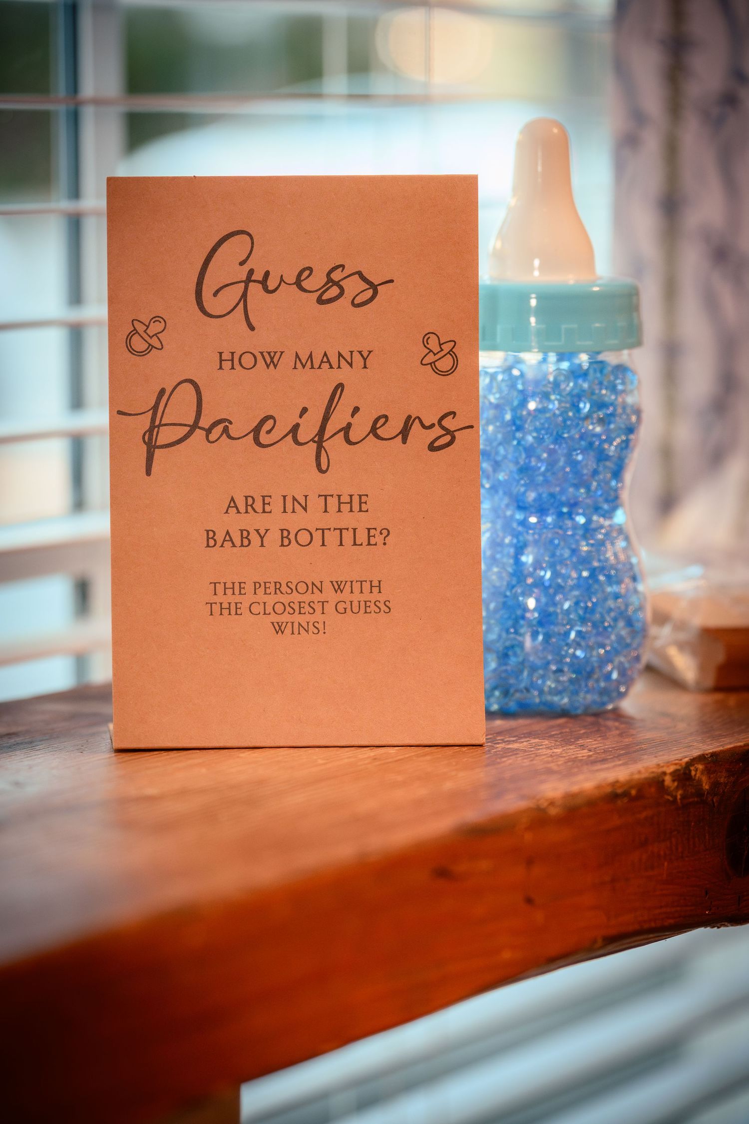 Blue candy pieces fill a decorative baby bottle next to brown cards with guessing game questions at a baby shower.