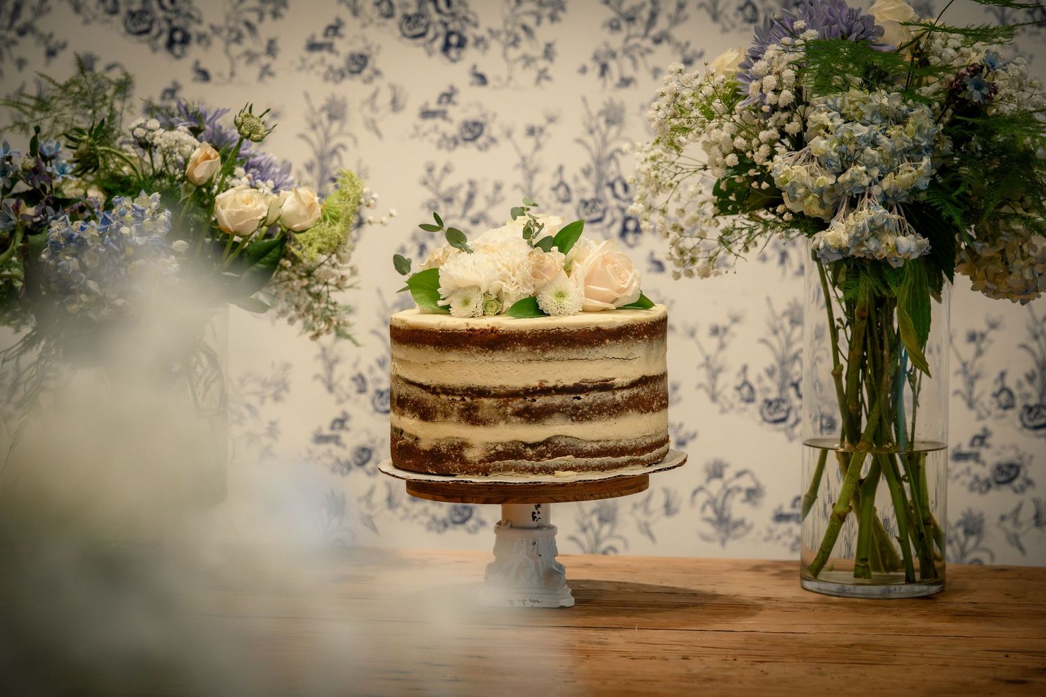 A rustic wedding cake with textured buttercream frosting and fresh flowers sits on a wooden table against floral wallpaper.