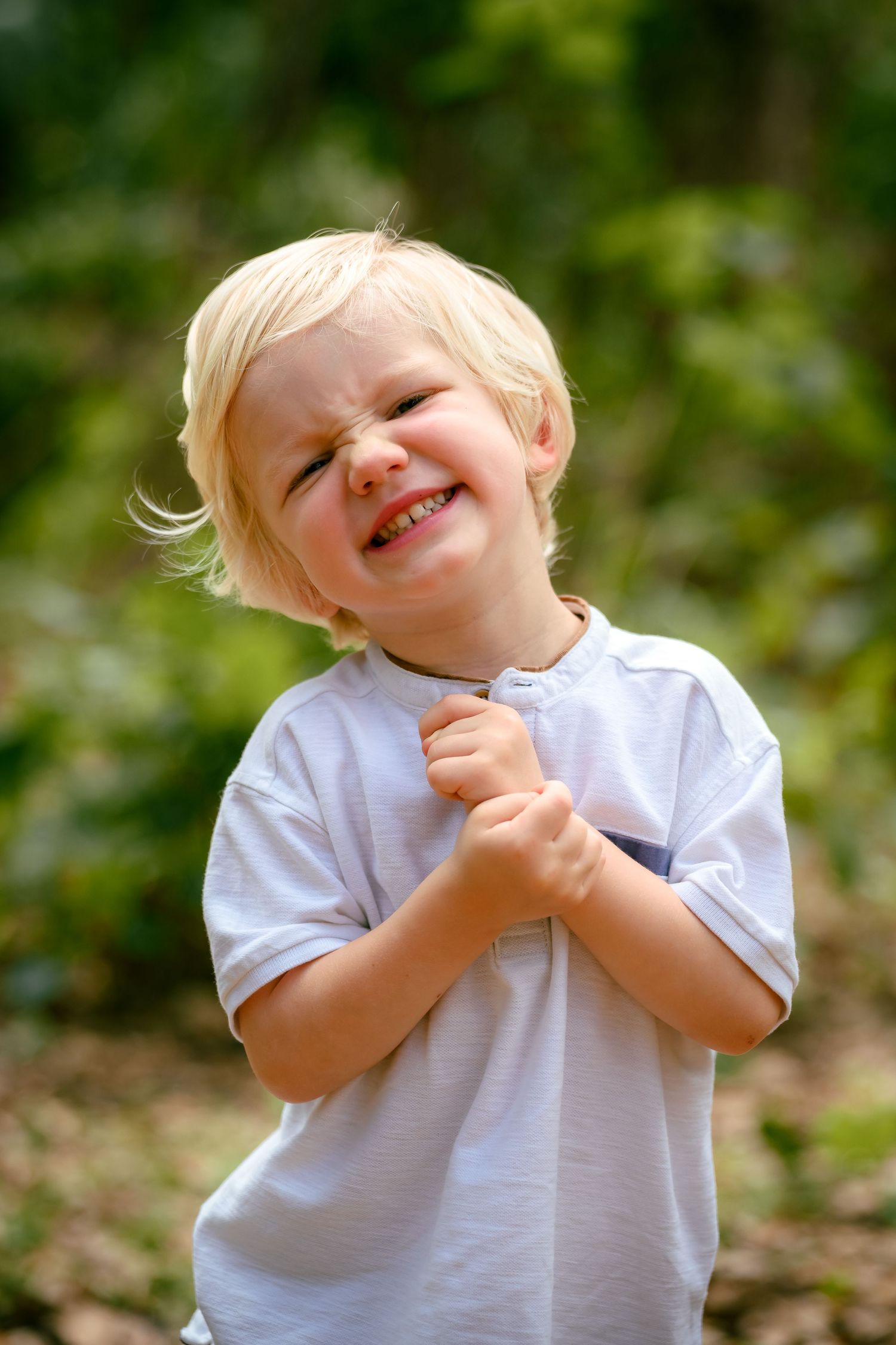 Cute blonde toddler boy squinting and smiling with clasped hands outdoors in a green park setting.