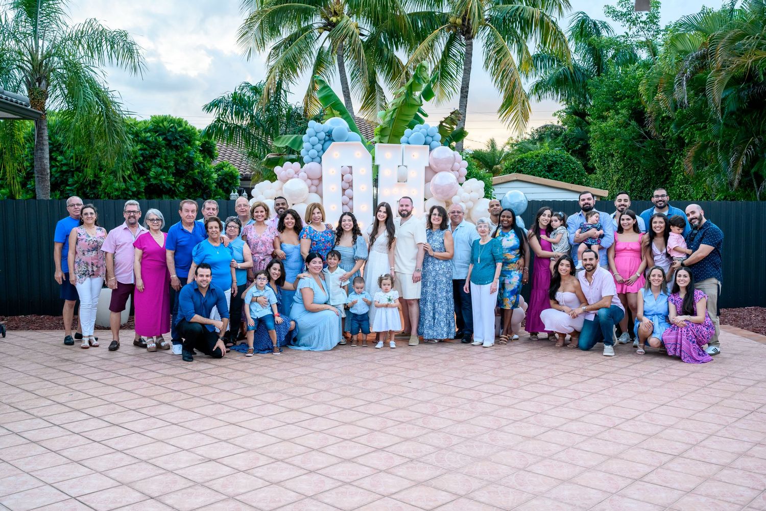 Large family group photo at an outdoor celebration with decorative backdrop, tropical greenery, and festive blue and pink decor.