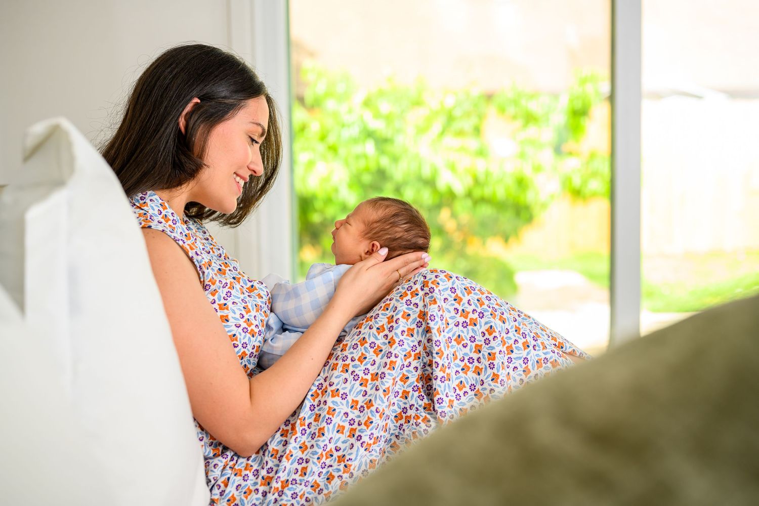 Happy mother sitting and cradling her sleeping newborn baby in a floral dress near a window.