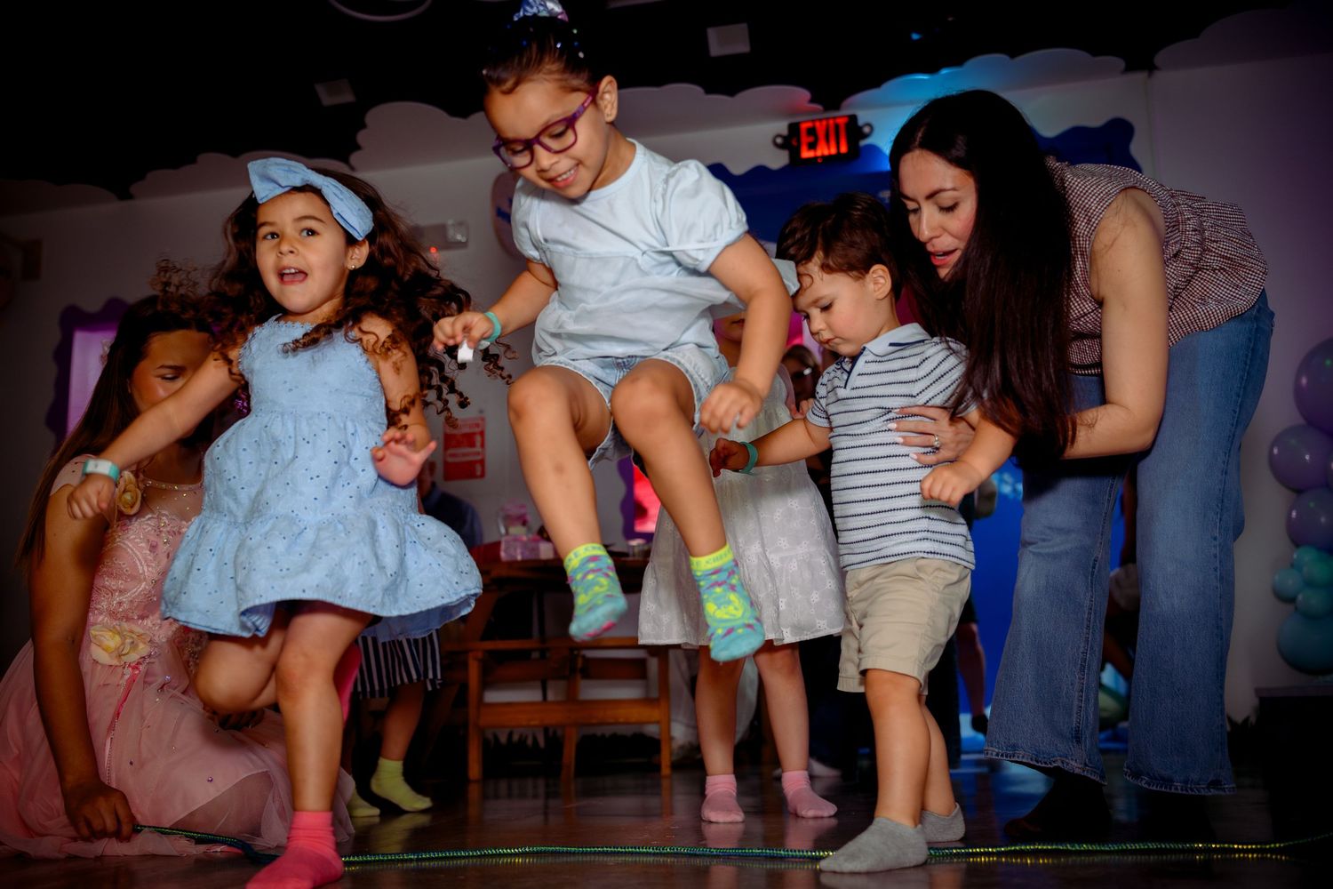 Children dancing and jumping joyfully at a colorful indoor birthday party with an adult supervising.