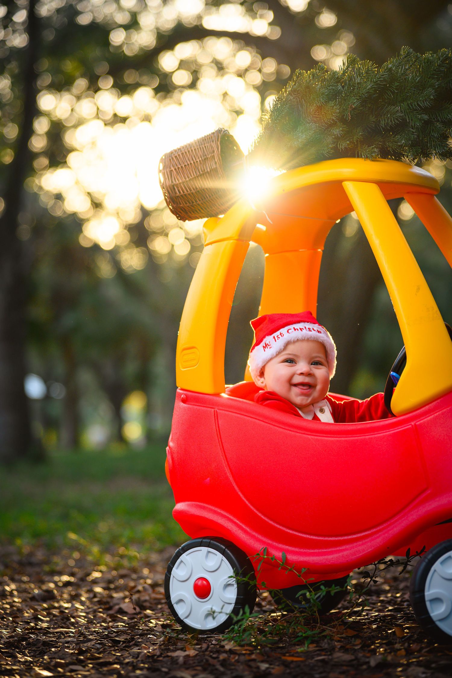 Laughing baby in Santa hat rides red and yellow toy car in sunlit park.