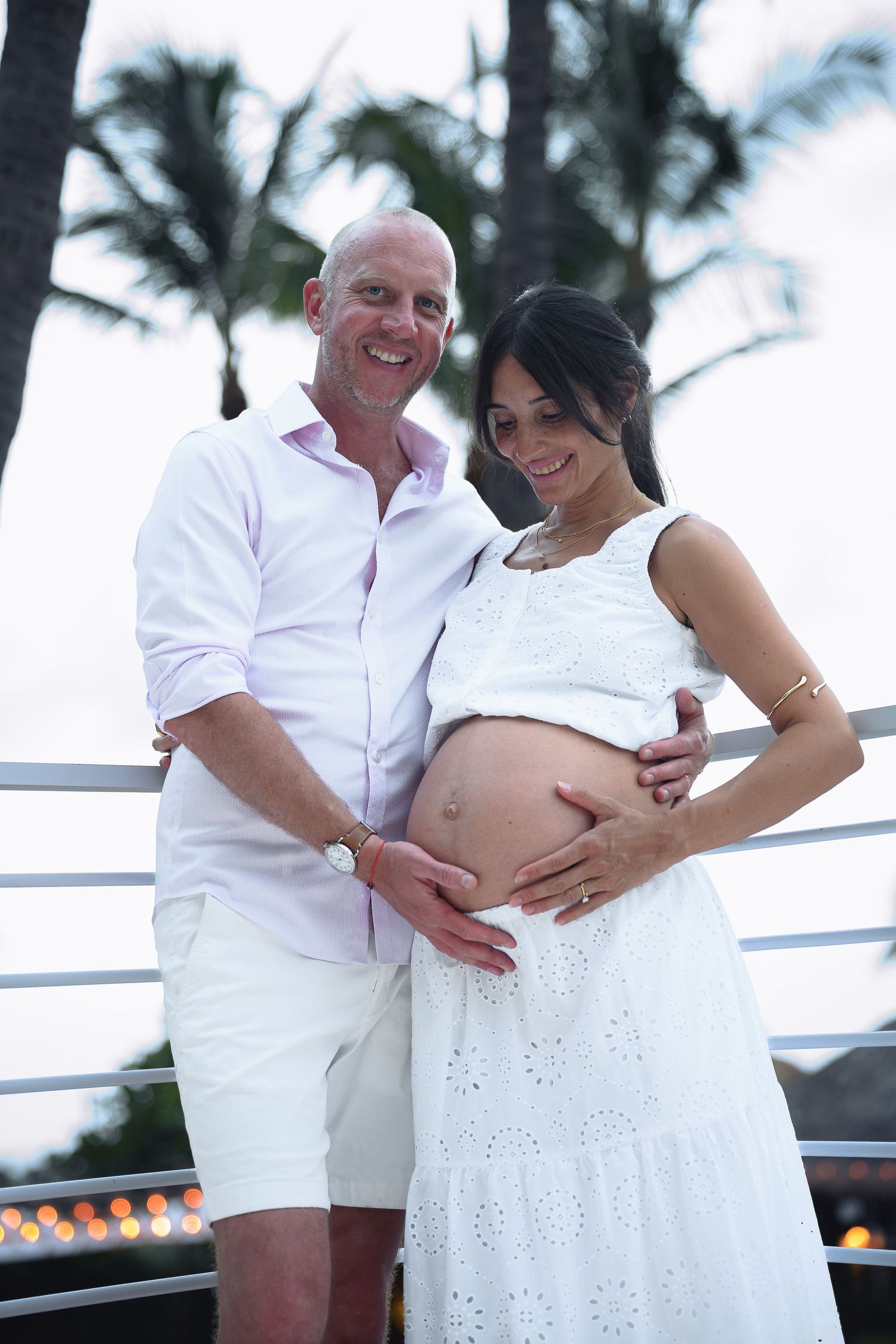Smiling couple in white outfits posing outdoors, man holding pregnant womans bare belly with palm trees behind.