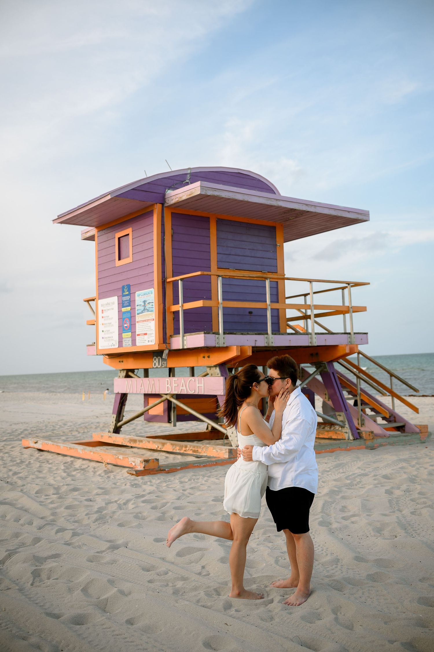 Couple kissing on Miami Beach sand in front of colorful purple and orange lifeguard tower.