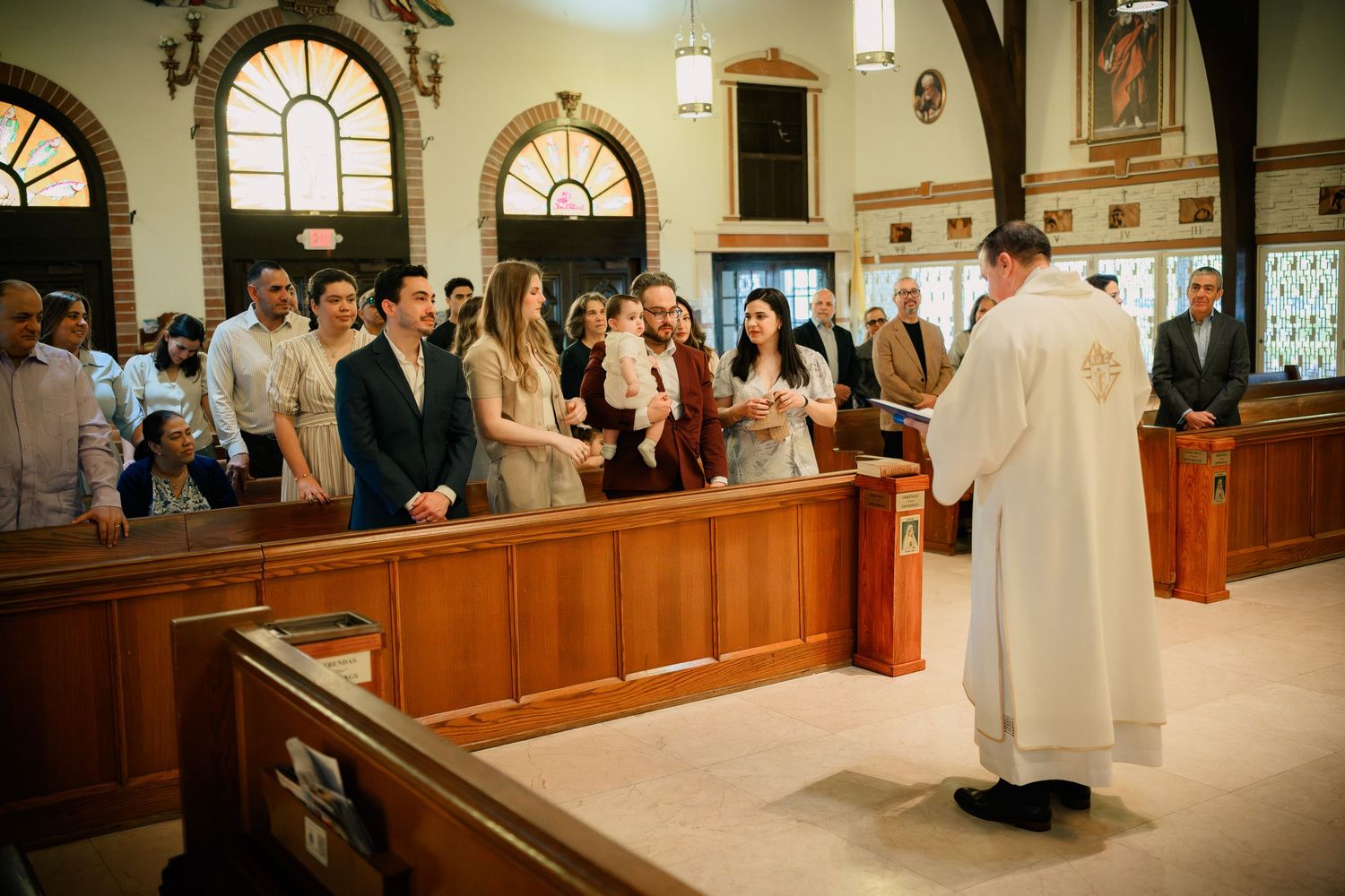 Priest officiating a Catholic church ceremony with young adults standing at the altar in a wooden pew sanctuary.