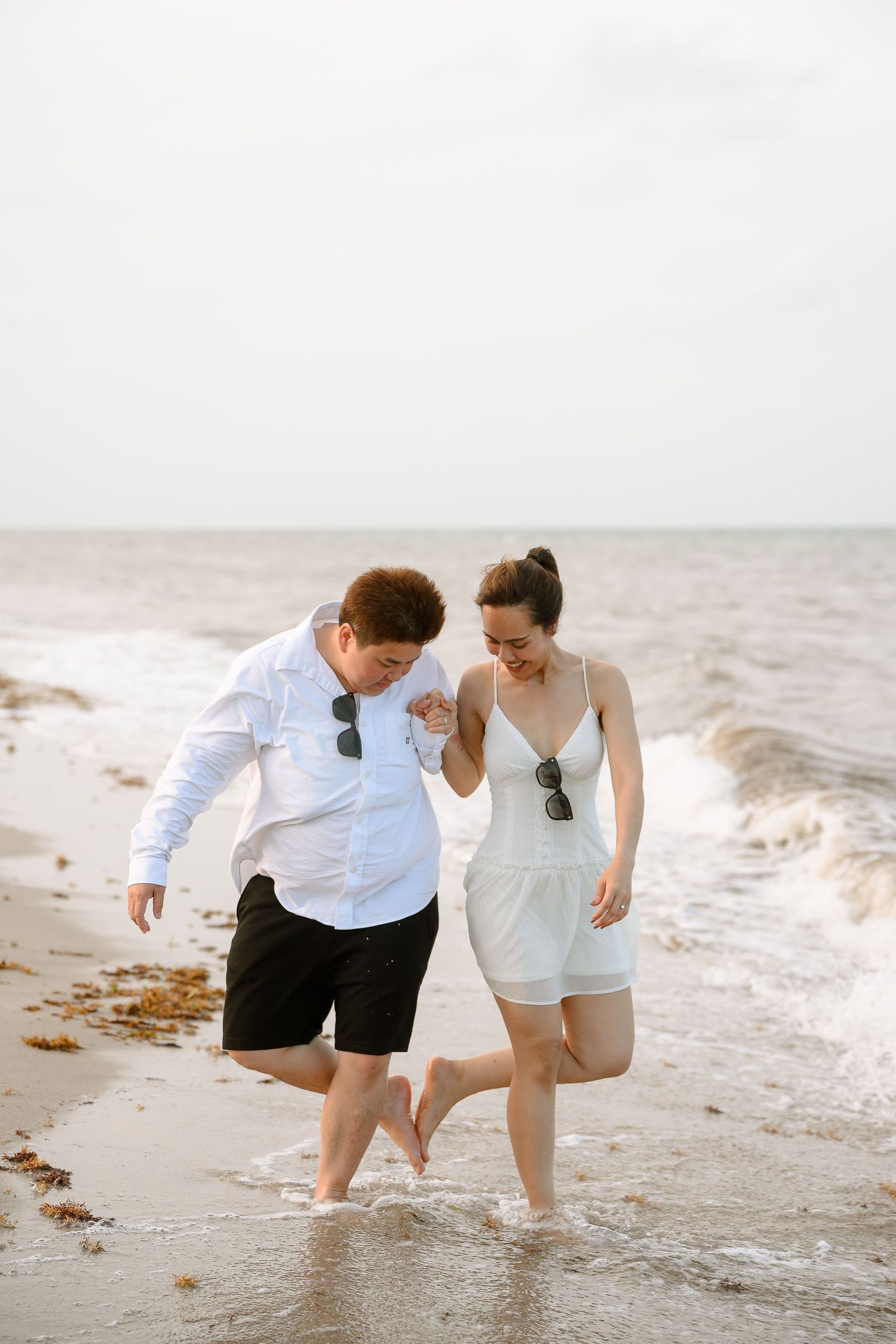 Couple laughing and splashing in ocean waves on a beach, dressed in white outfits with sunglasses.