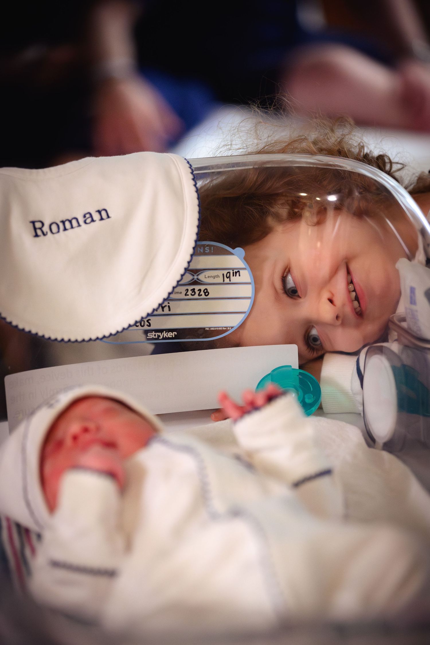 Curious toddler peering into hospital bassinet to look at newborn baby named Roman.
